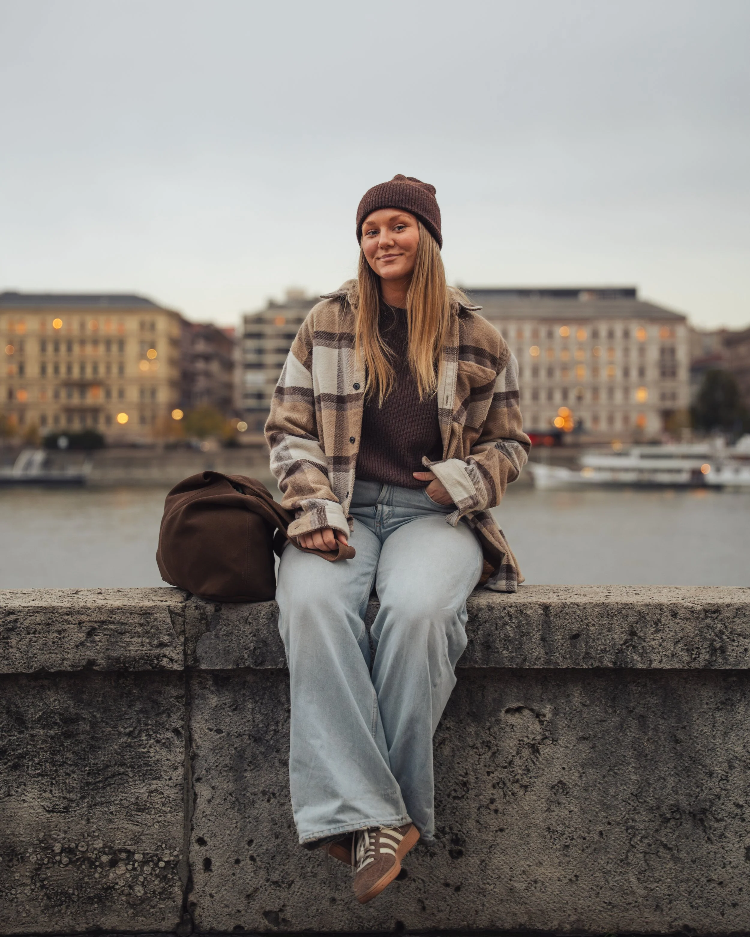 A young woman wearing a brown beanie, plaid jacket, and jeans sitting on a stone ledge by the water with buildings in the background during dusk.
