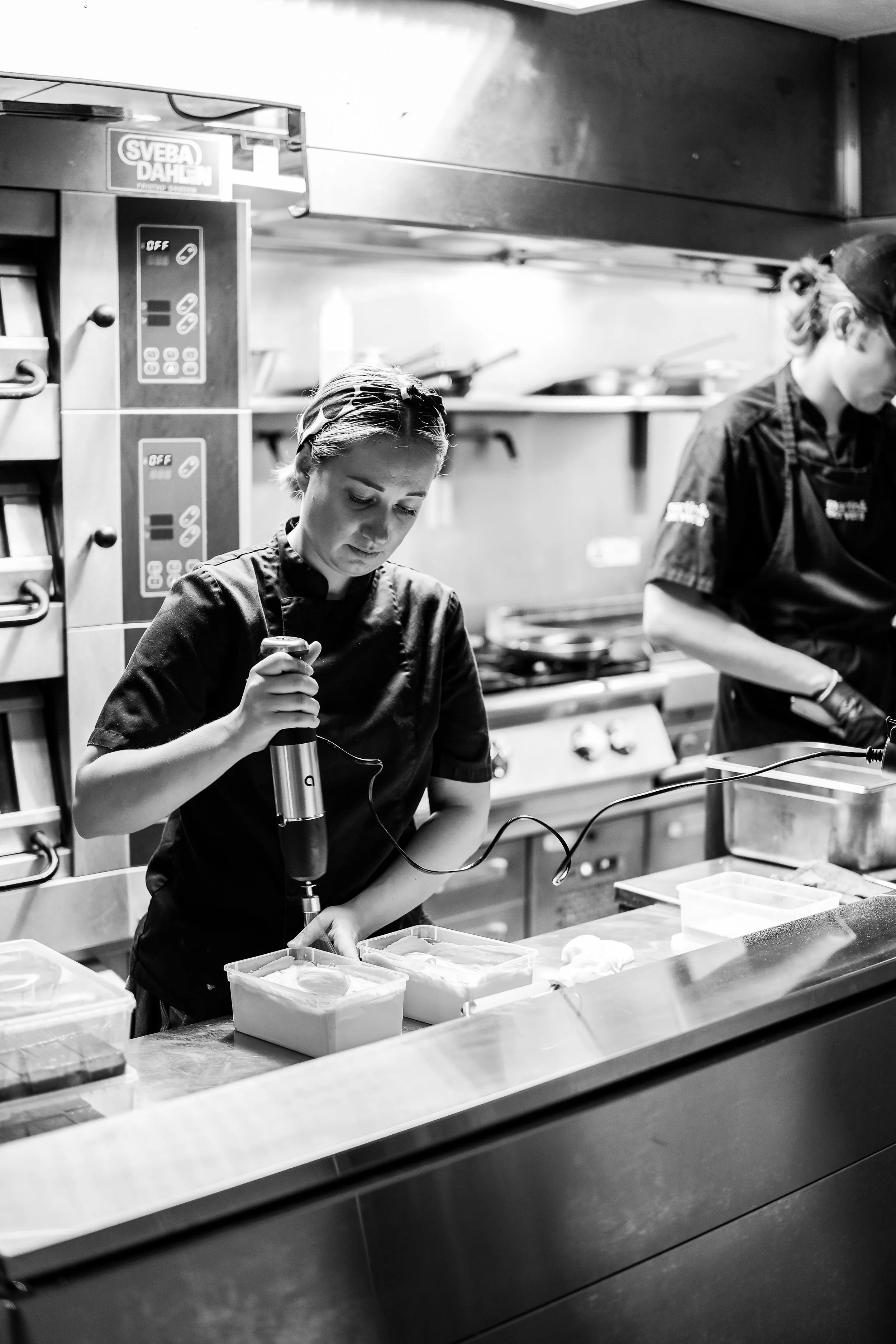 Chef preparing food in a professional kitchen with another chef visible in the background, breaking in a chef's coat