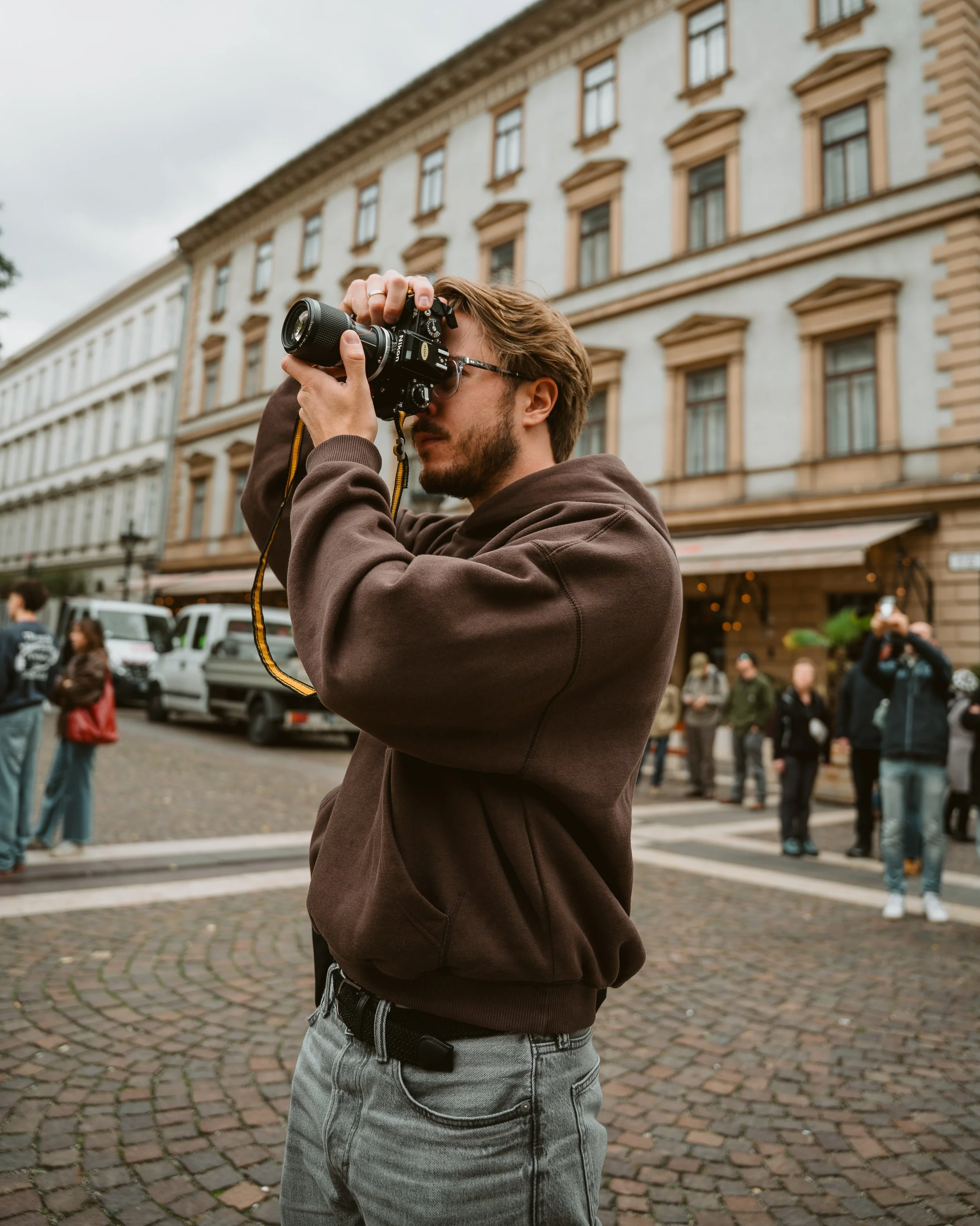 A man with brown hair, glasses, and a beard taking a photo with a camera on a city street with old buildings and people in the background.