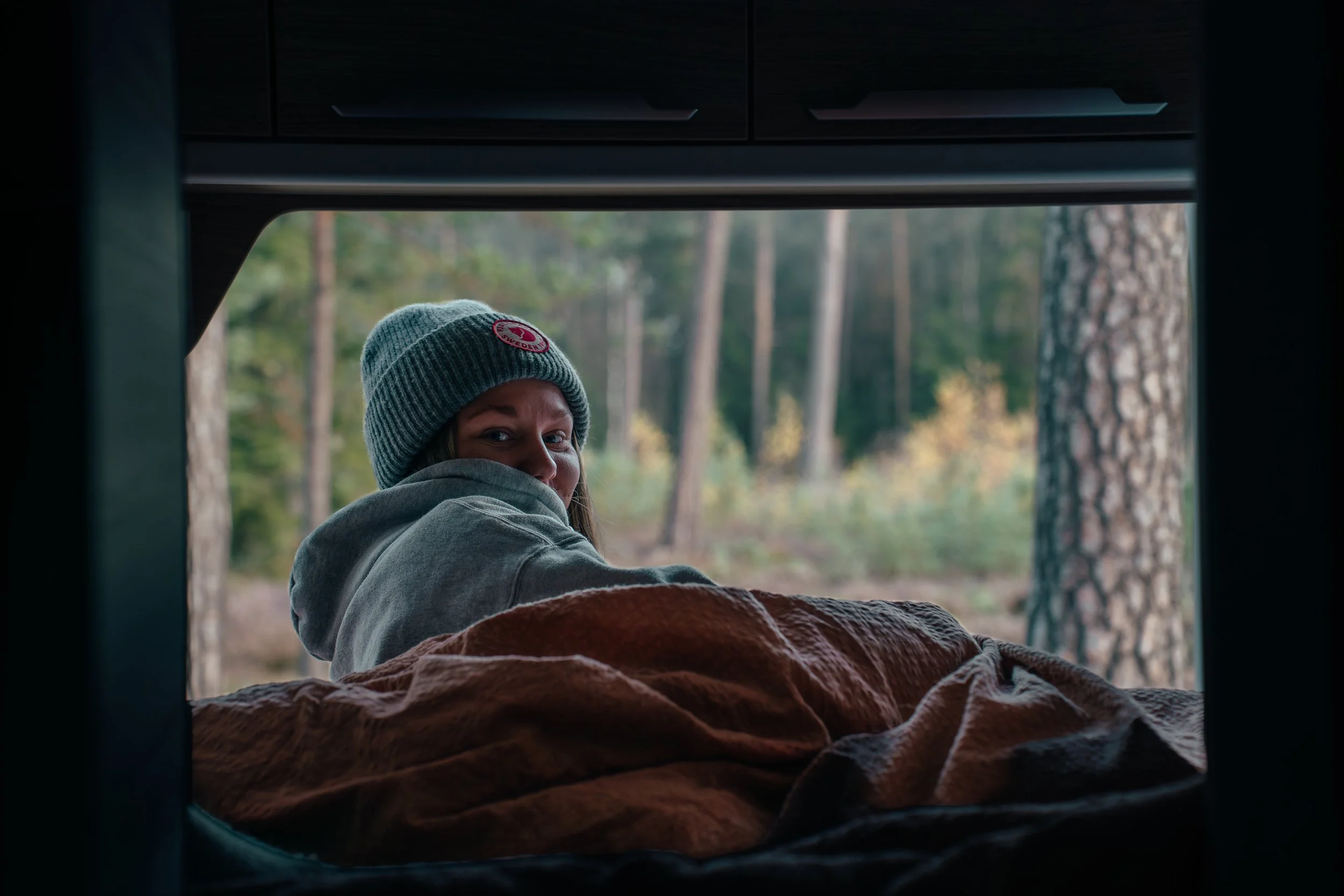Woman lying inside a camper, looking out at a forest with trees, wearing a gray hoodie and a knitted beanie.