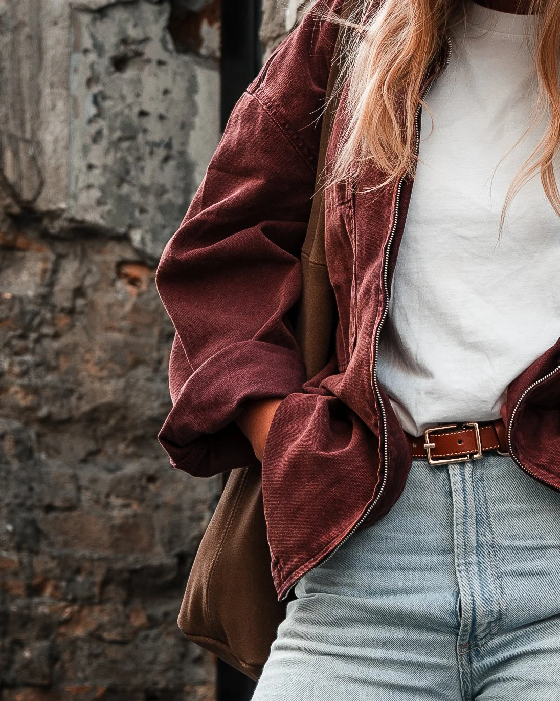 Close-up of a person wearing a white T-shirt, a maroon jacket, light blue jeans, and a brown belt, with a stone wall in the background.