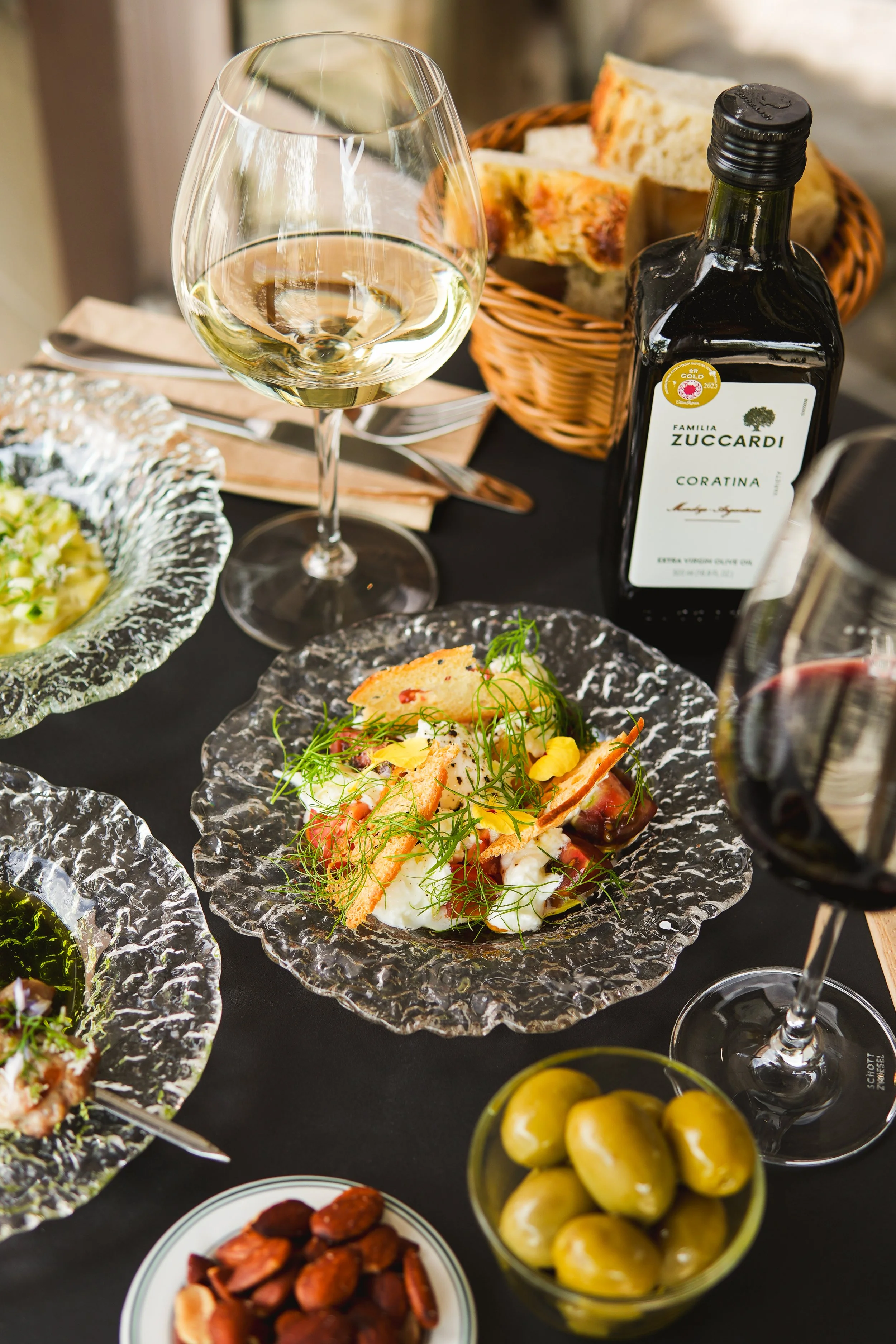 A dining table with a glass of white wine, a glass of red wine, a bottle of Zuccardi Coratina olive oil, a basket of bread, a plate of salad, a bowl of green olives, a small bowl of nuts, and dishes of other appetizers on a dark tablecloth.