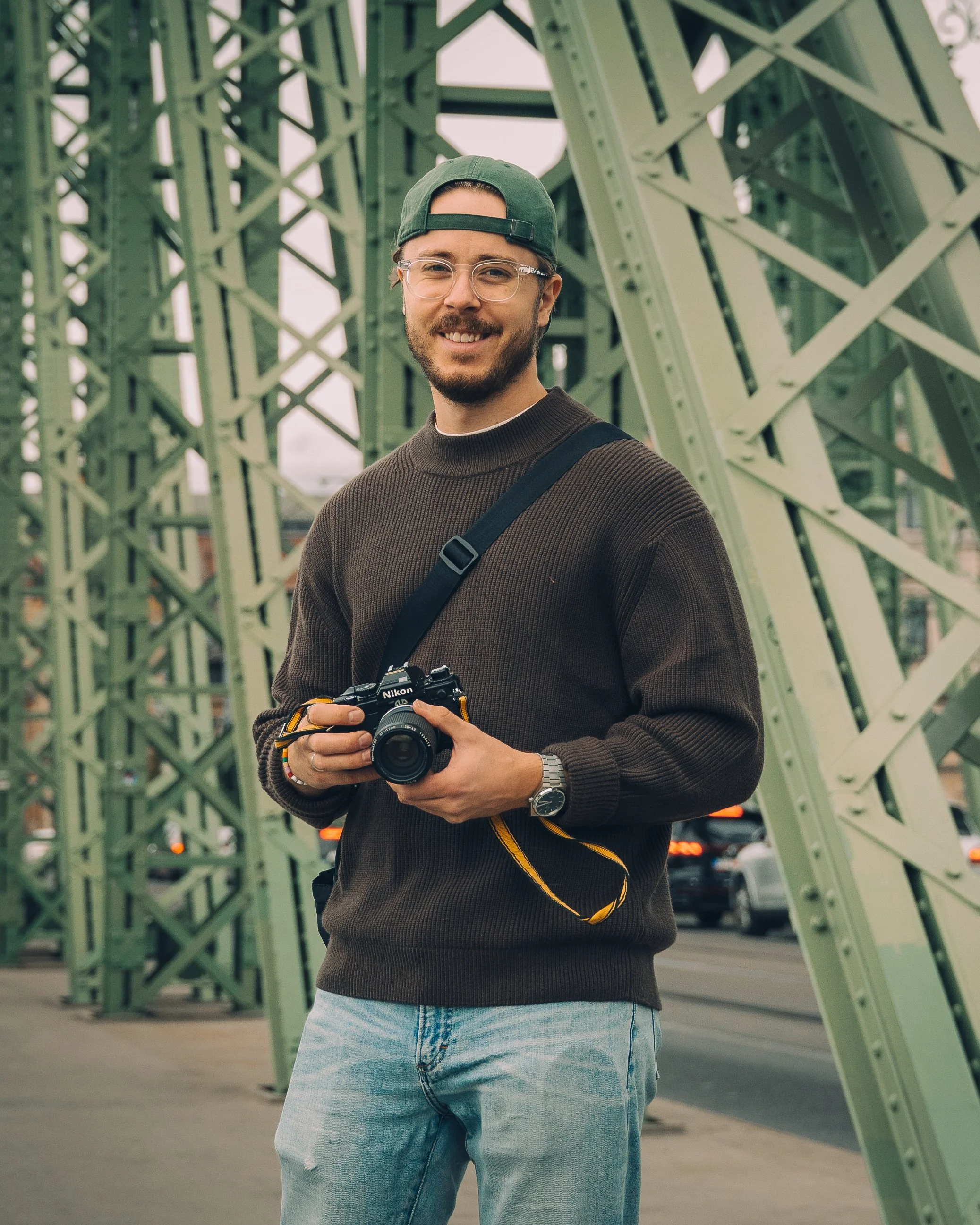 Young man with glasses and a beard smiling while holding a camera outdoors on a bridge with green metal beams.