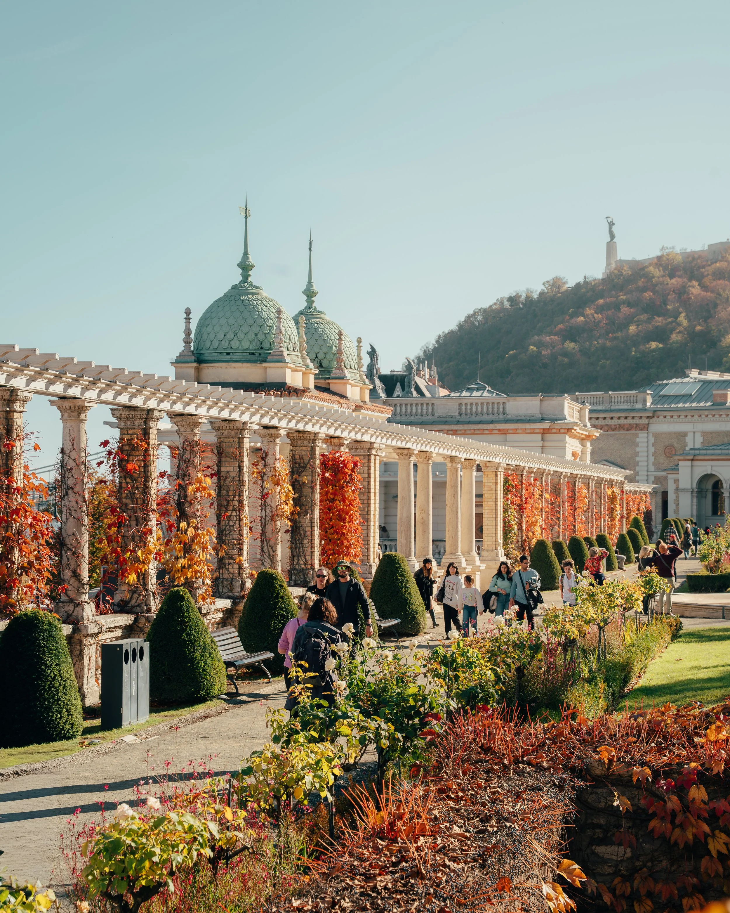 People walking through a garden with trimmed bushes and colored autumn leaves, next to a building with domed towers and columns, with a hill and statue in the background.