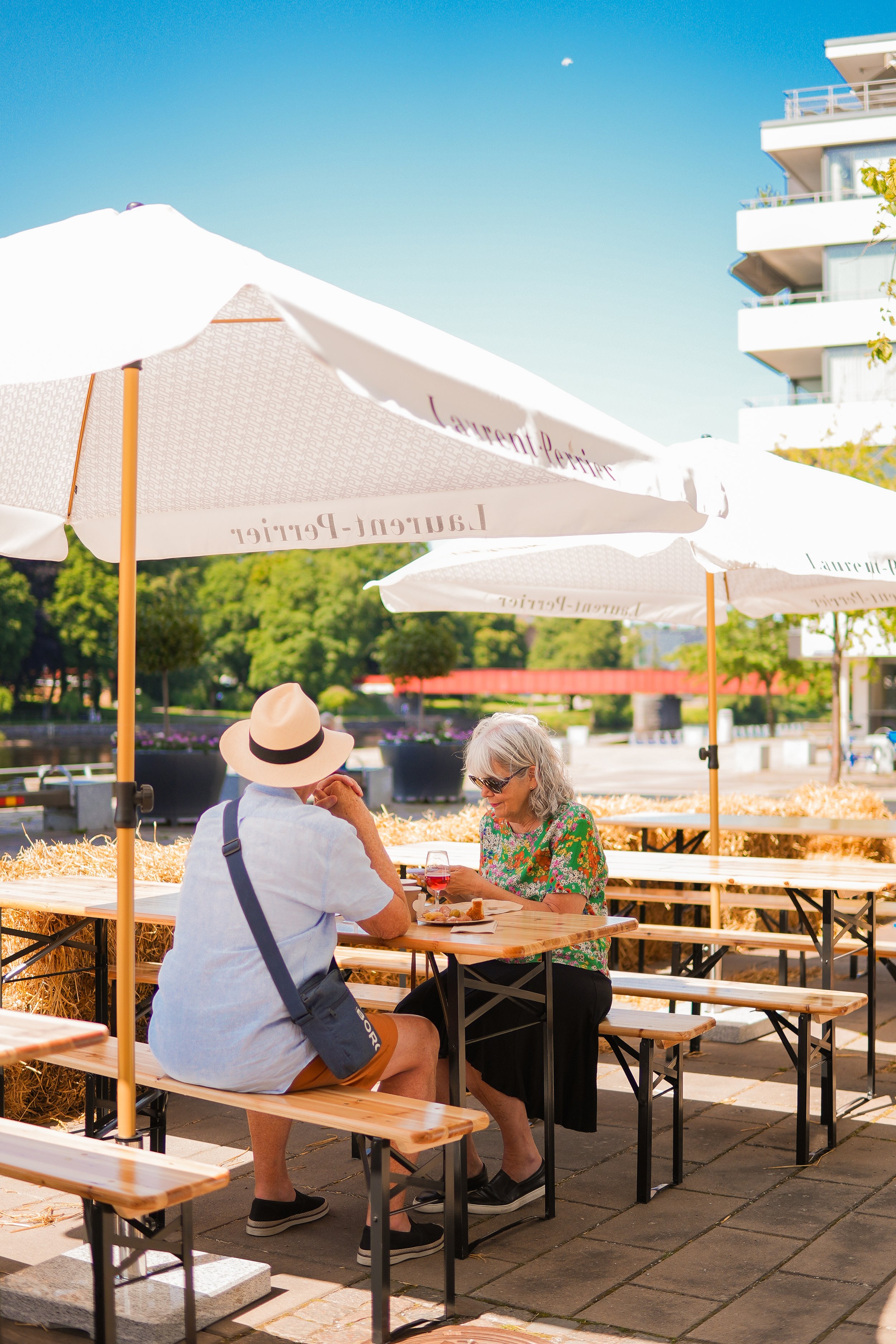 An elderly woman and a man sitting at an outdoor table under umbrellas, enjoying a meal and drinks on a sunny day.