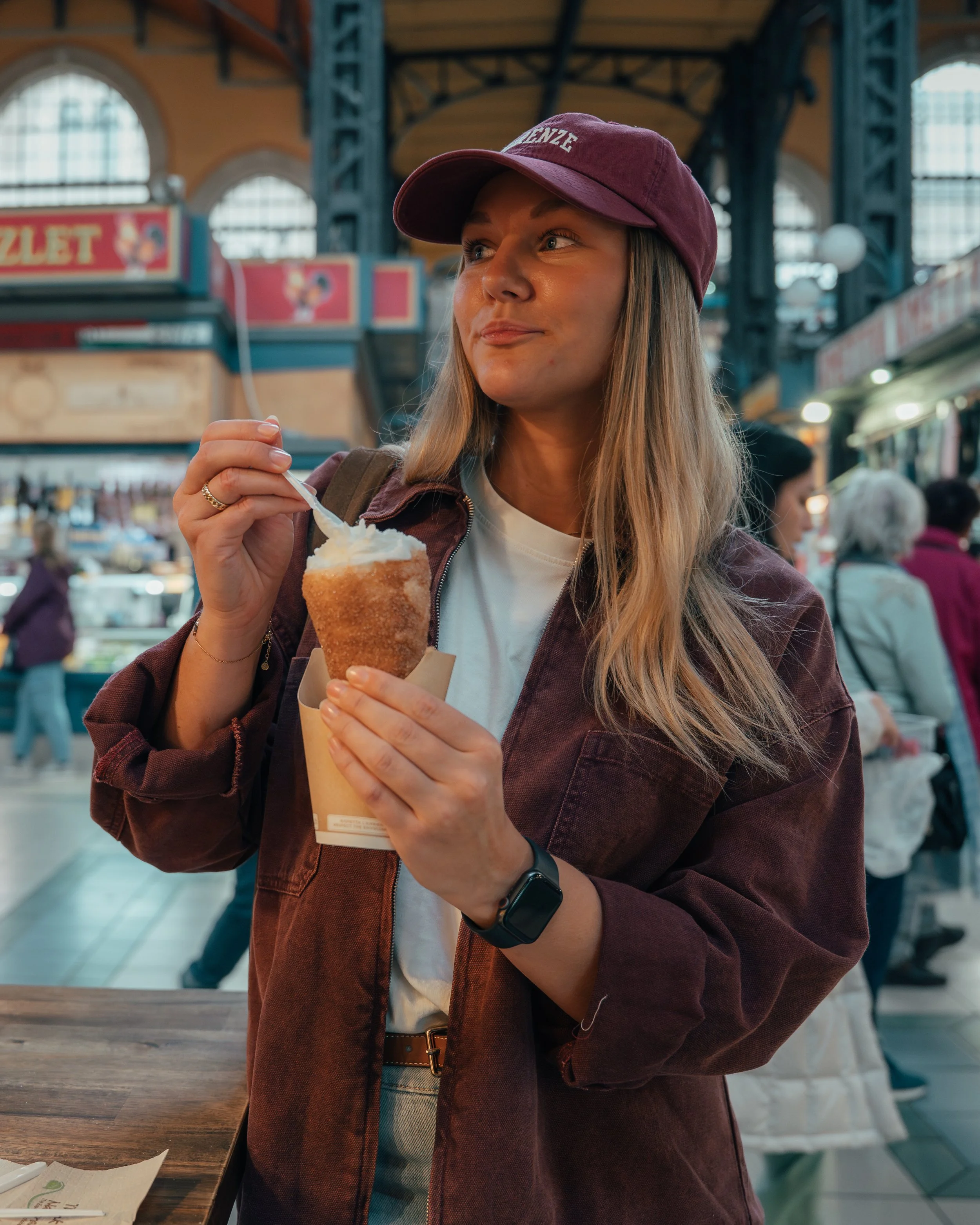 A woman wearing a maroon cap and jacket enjoying an ice cream cone at an indoor market.