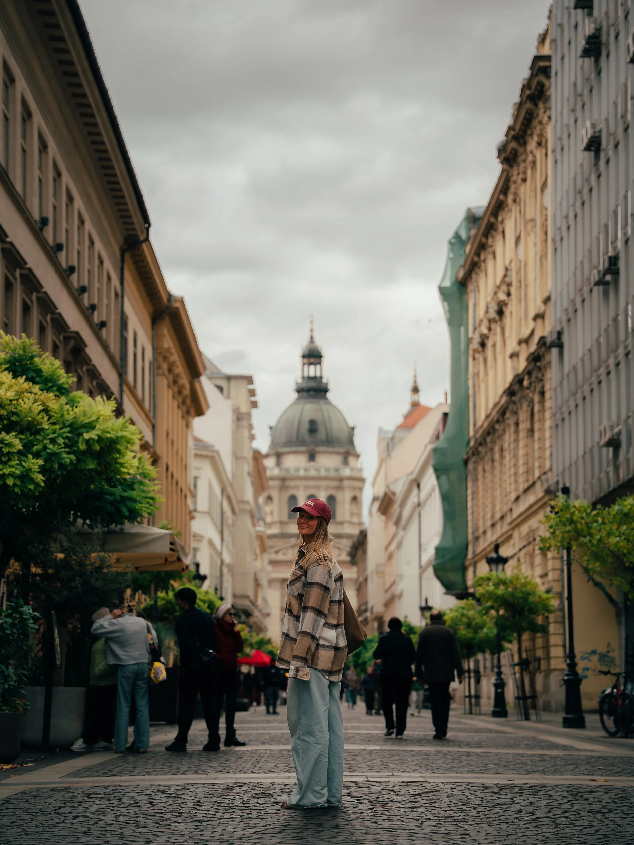 A young woman in a plaid jacket and gray pants standing on a cobblestone street, with historic buildings and a large domed building in the background, under a cloudy sky.