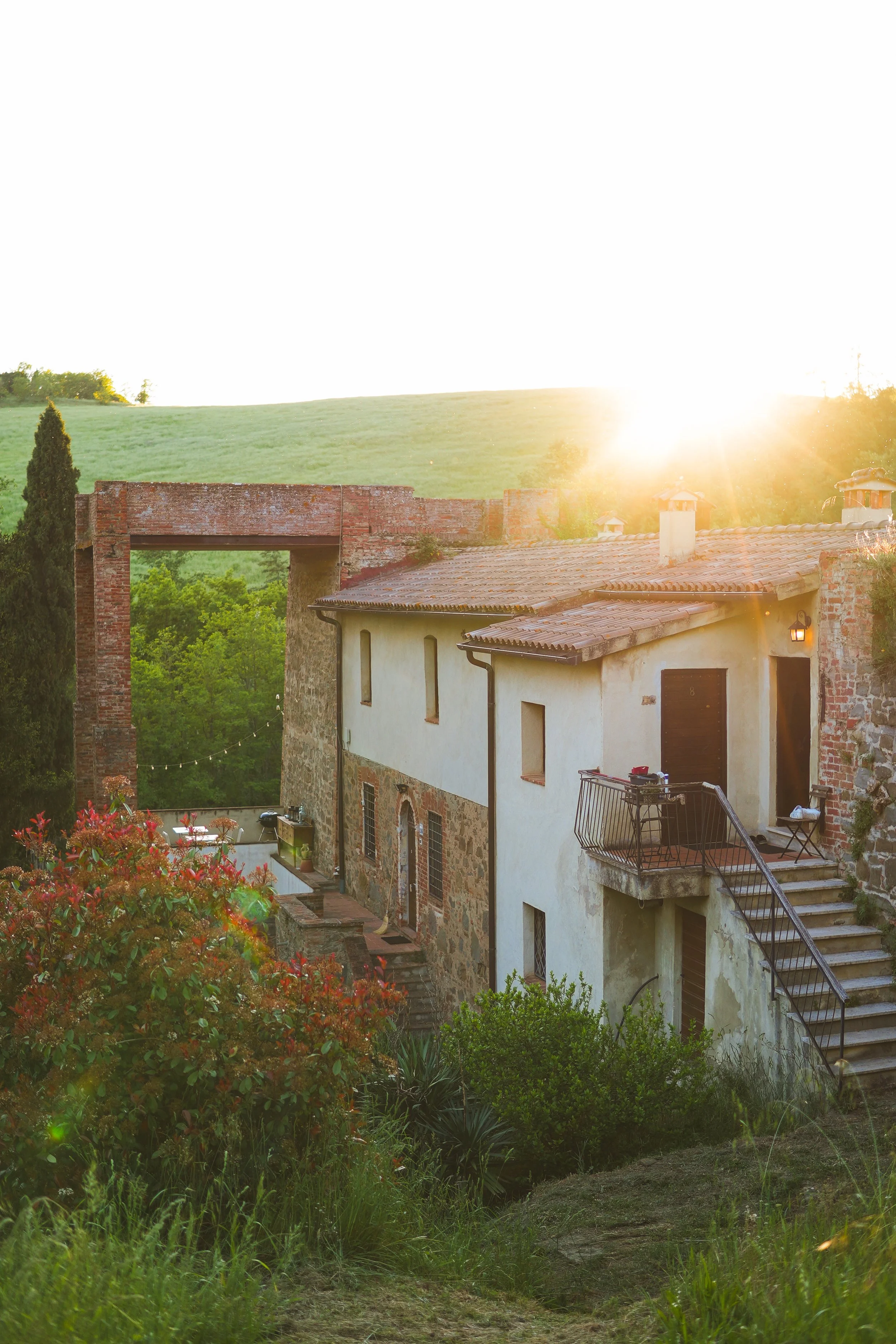 A rustic house with a tiled roof and a small balcony, set in lush greenery, with the sun setting behind a hill in the background.