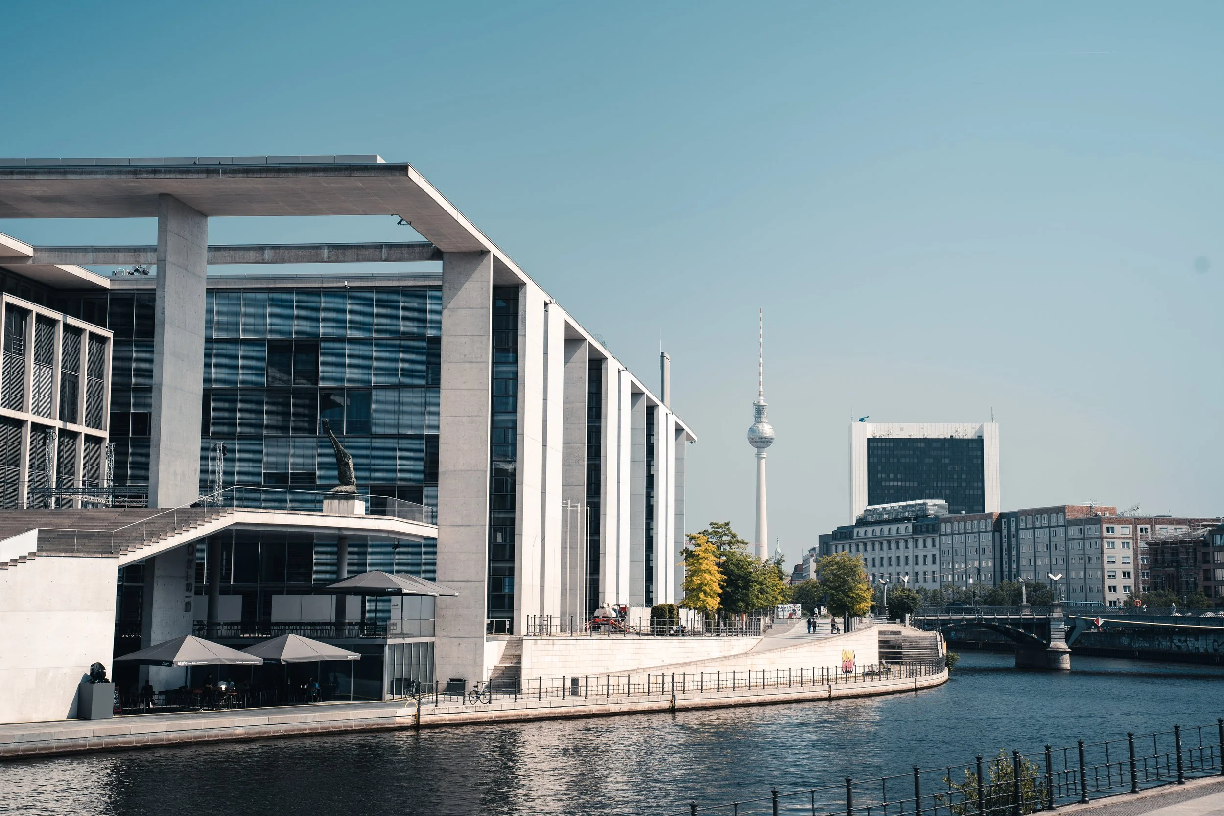 Modern cityscape with contemporary buildings along a river, featuring a bridge and a television tower in the background, under a clear blue sky.