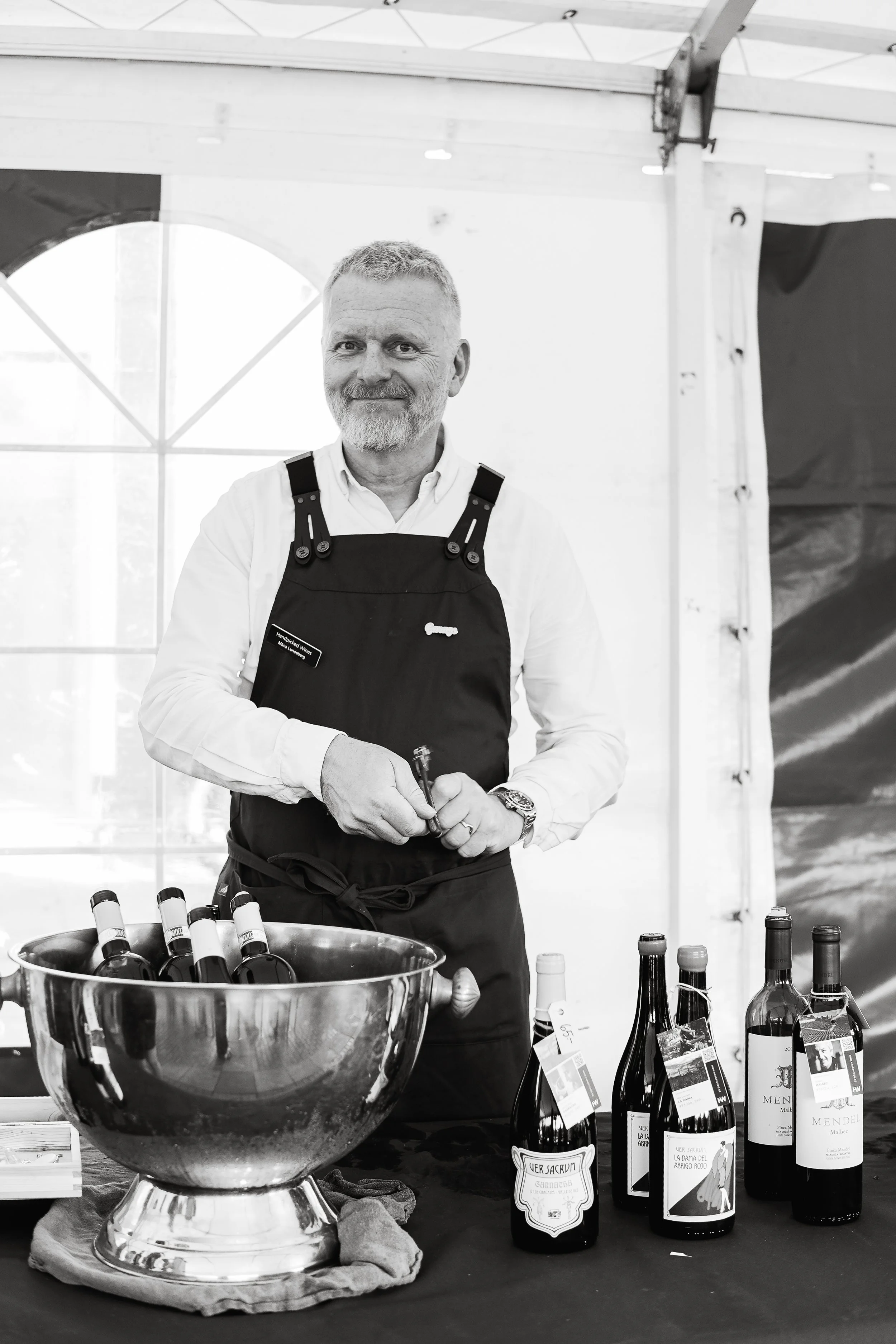 A man in a white shirt and black apron stands behind a table with wine bottles at a wine tasting event inside a tent.