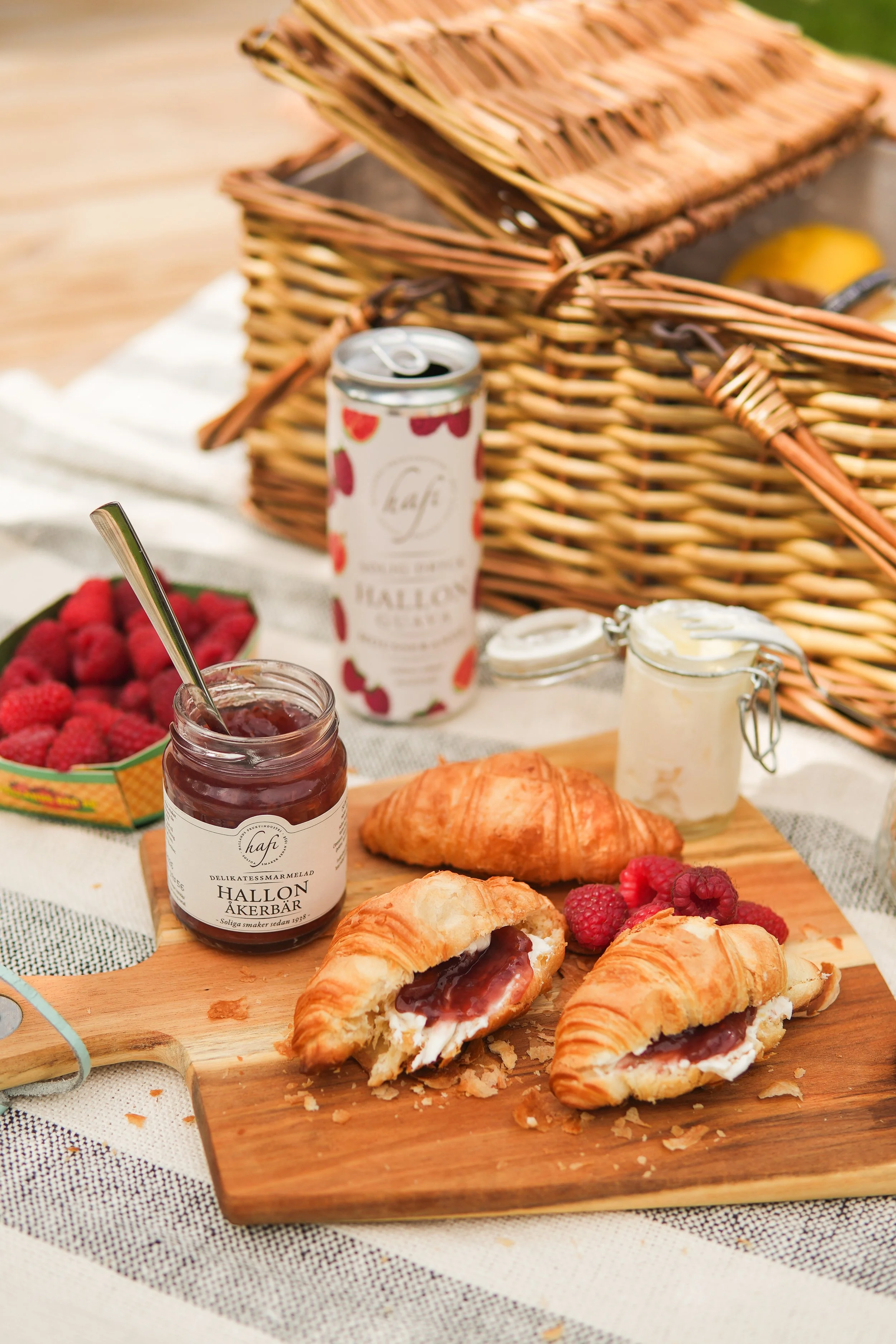 Close-up of a picnic table with croissants filled with jam and cream, fresh raspberries, a jar of raspberry jam, a can of soda, and wicker baskets in the background.