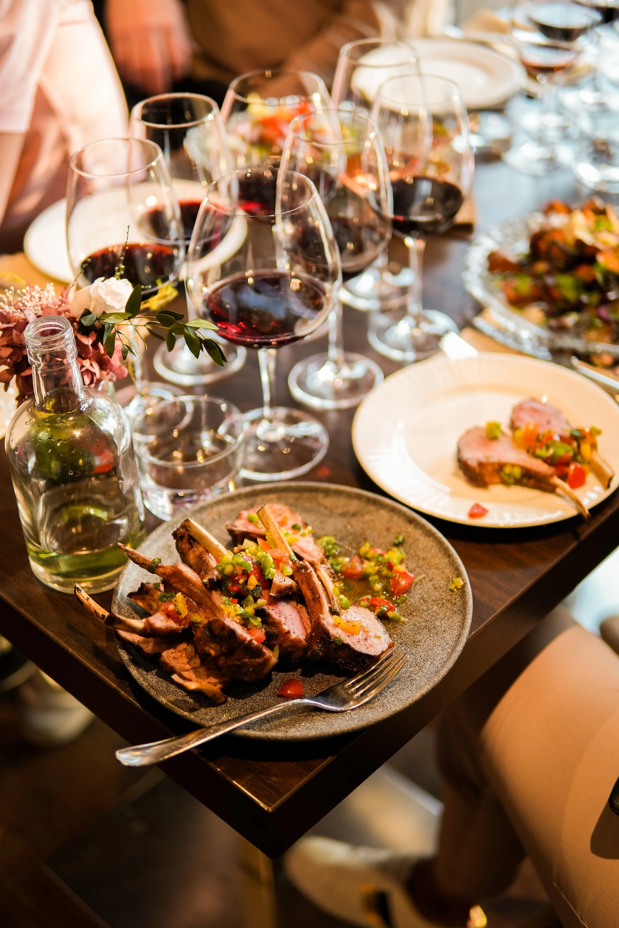 A dinner table with multiple glasses of red wine, plates of grilled meat with vegetable toppings, and a bottle of water, accompanied by a small vase of flowers.
