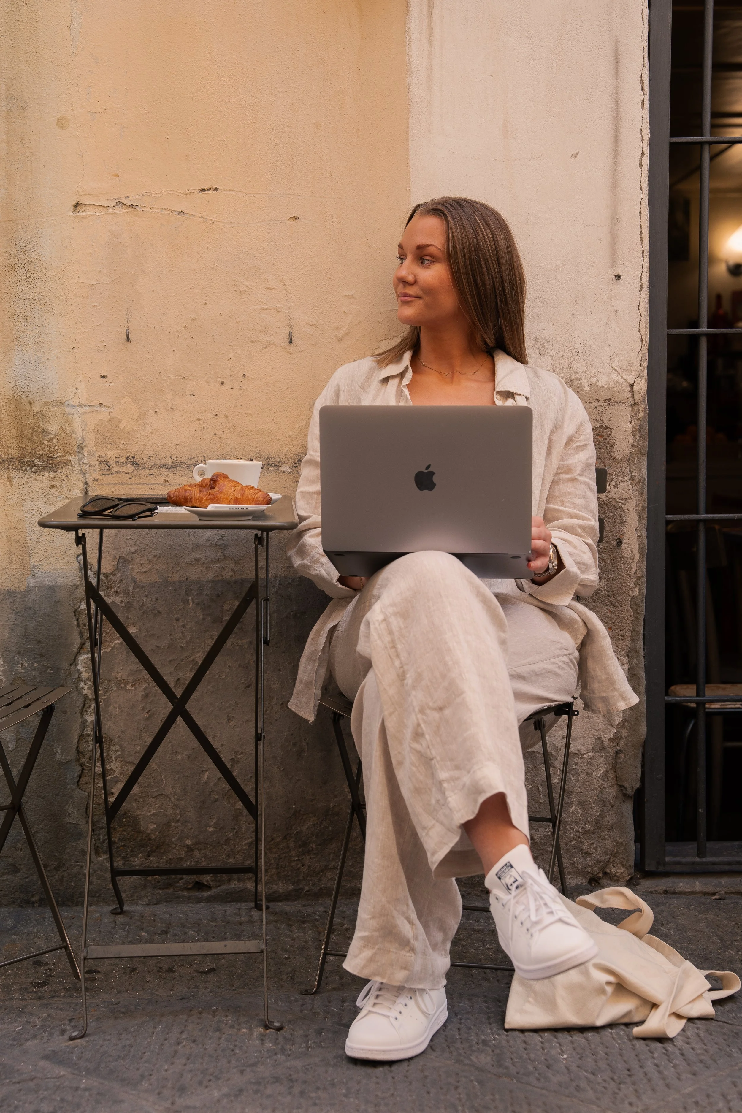 A woman sitting outdoors on a folding chair, holding a MacBook, with a small table beside her holding a croissant, coffee, and glasses, against an old textured wall.