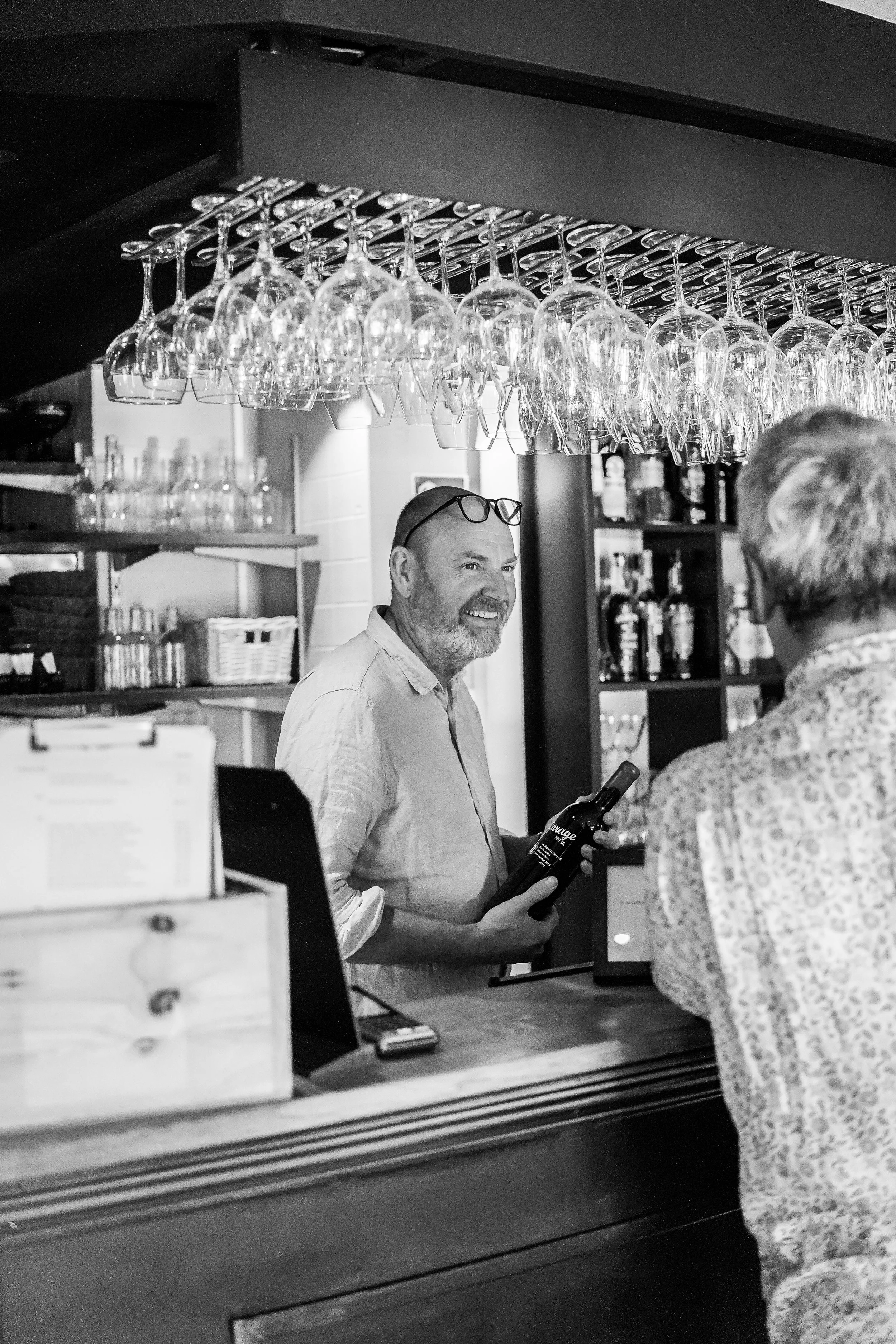A bartender with glasses on his head and a beard, smiling while holding a wine bottle, talking to a customer at a bar with glasses hanging above.