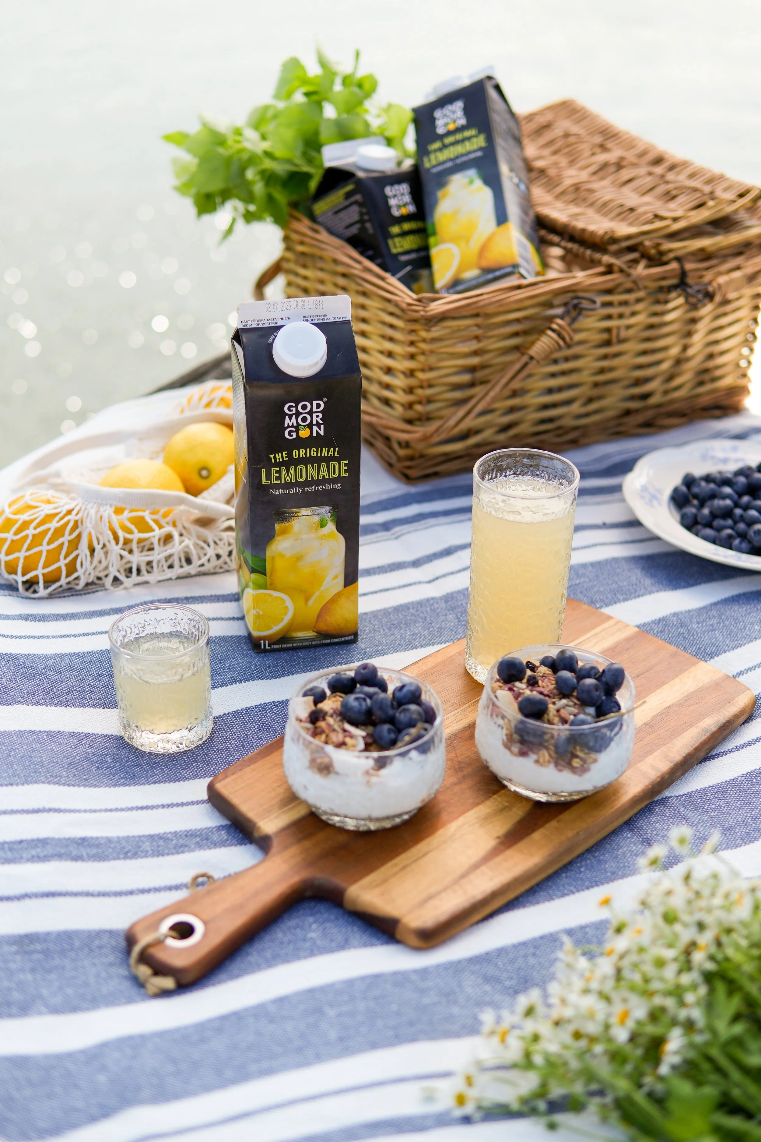A picnic scene with a striped blue and white tablecloth, featuring bowls of yogurt with blueberries and granola, glasses of lemonade, and a carton of God Morning lemonade. In the background, there's a wicker basket with more juice cartons, a bunch of