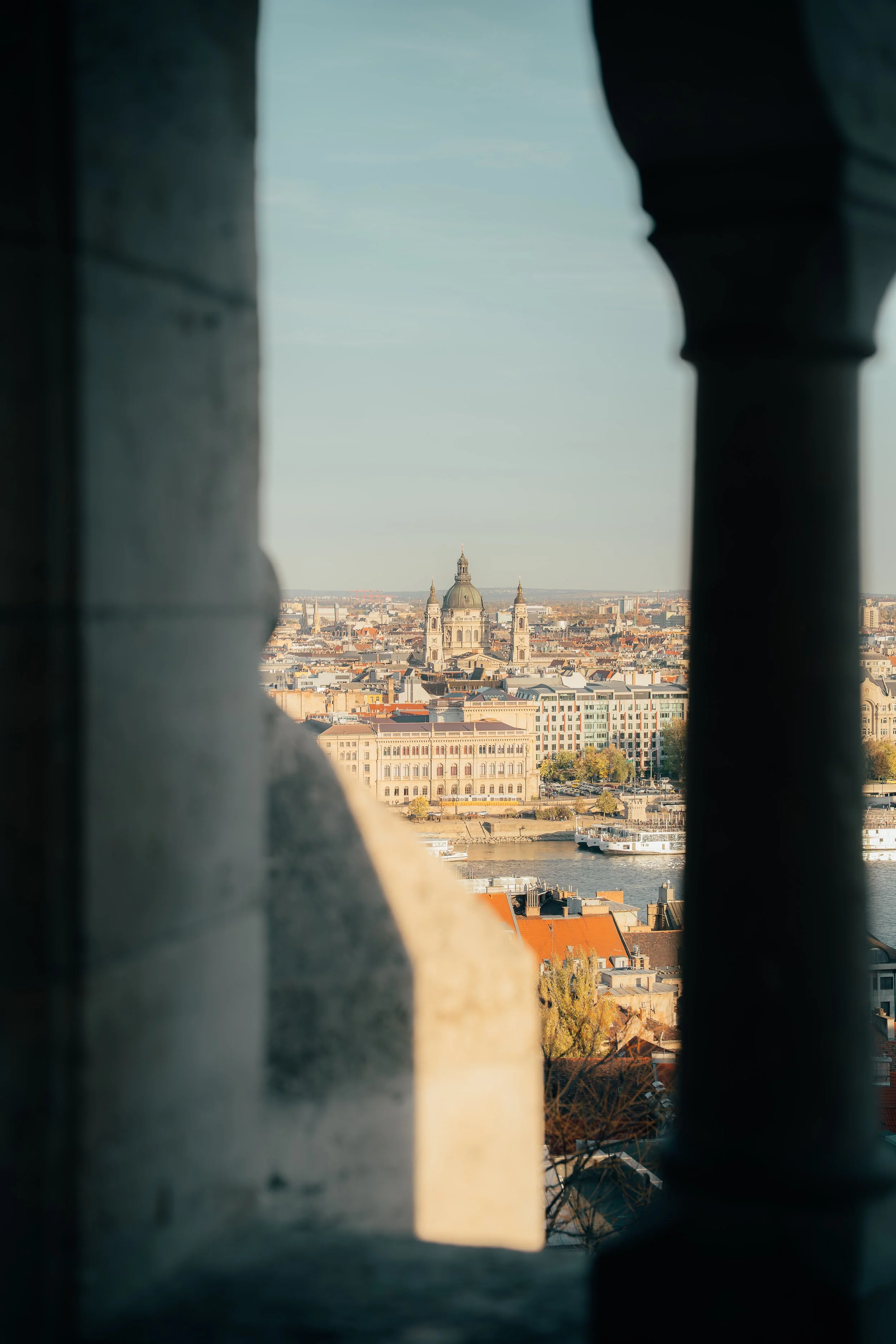 View of a city skyline with a river in the foreground, seen through a stone or concrete architectural opening with columns, featuring a prominent domed building with two towers in the background.