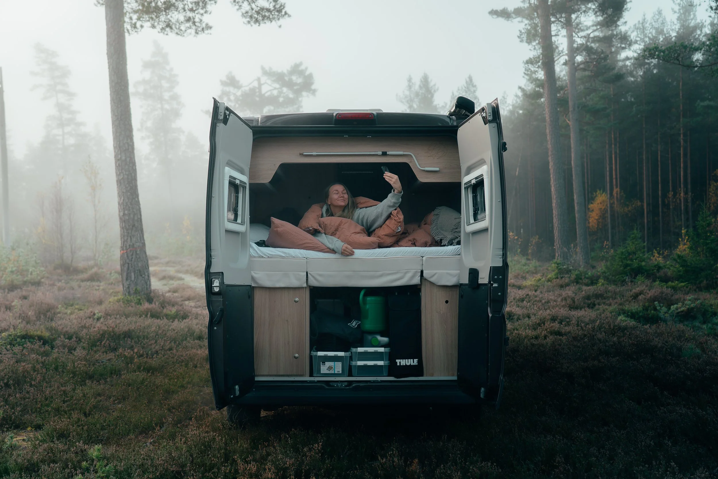 Woman lying in bed inside a camper van parked in a forest, taking a selfie.