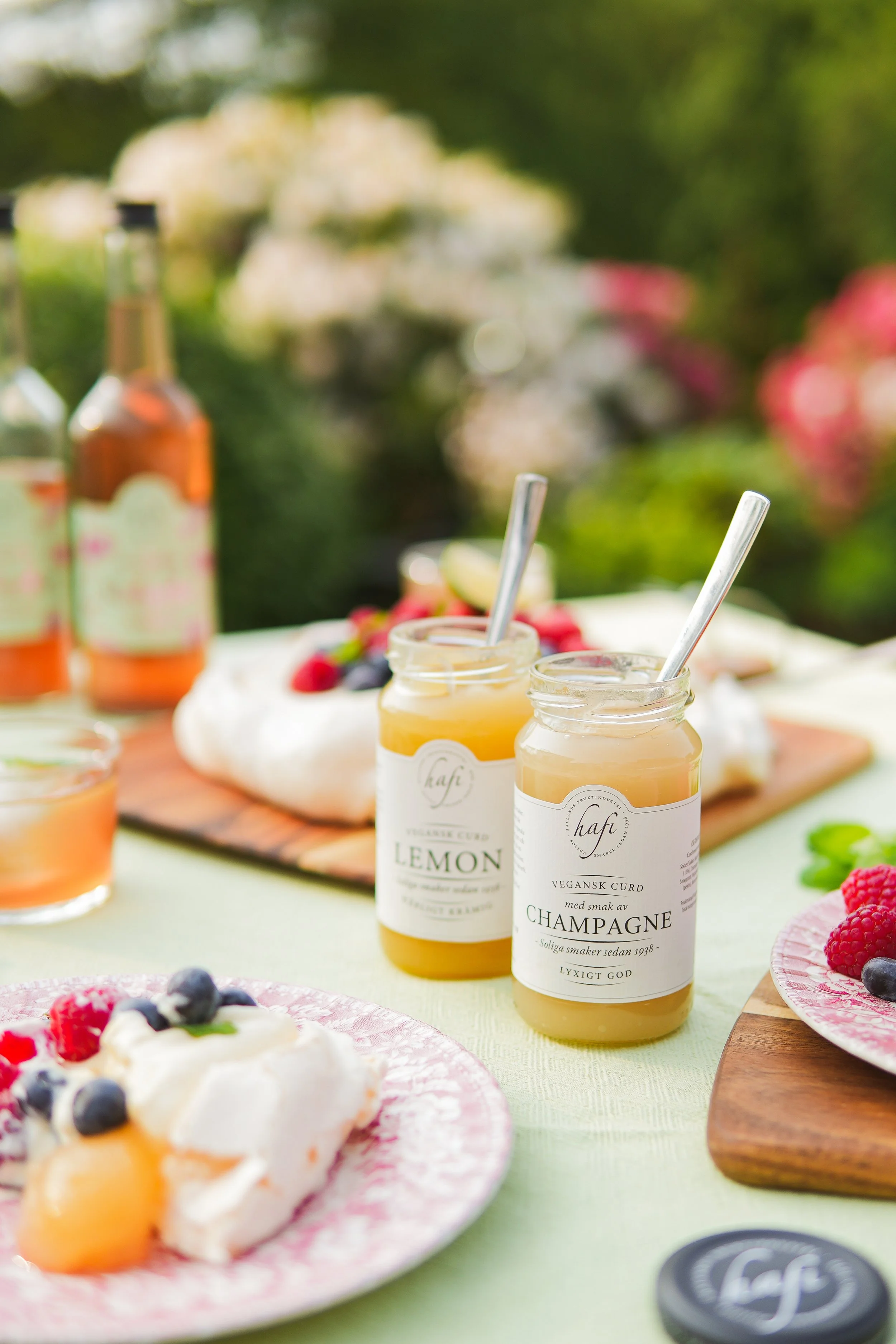 Outdoor table set with jars of lemon and champagne-flavored vegan curd, plates of cake with berries and fruit, and bottles of beverages in the background, surrounded by greenery and flowers.