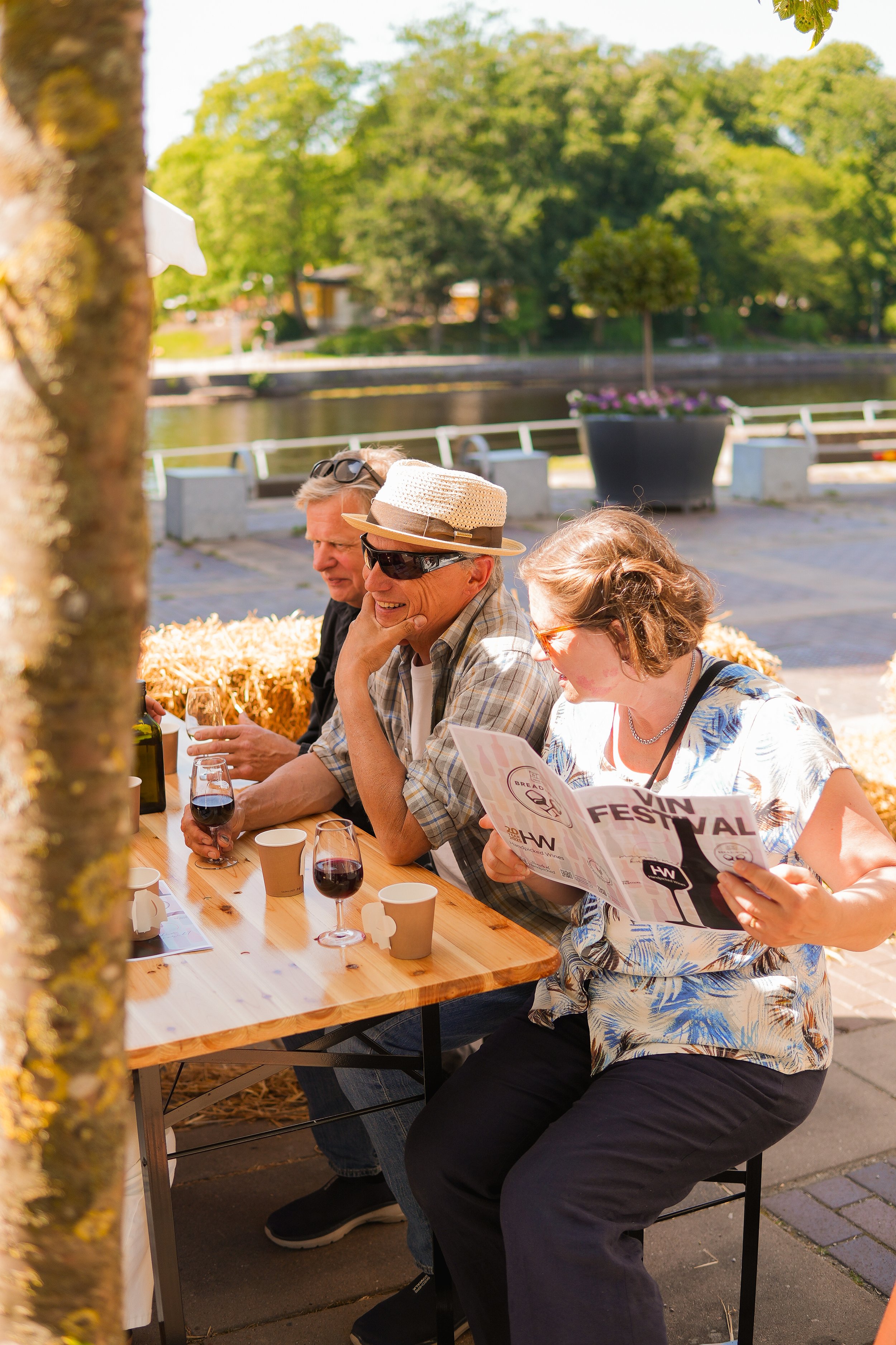 People sitting at a wooden table outdoors enjoying wine and reading a festival brochure, with trees and a river in the background.