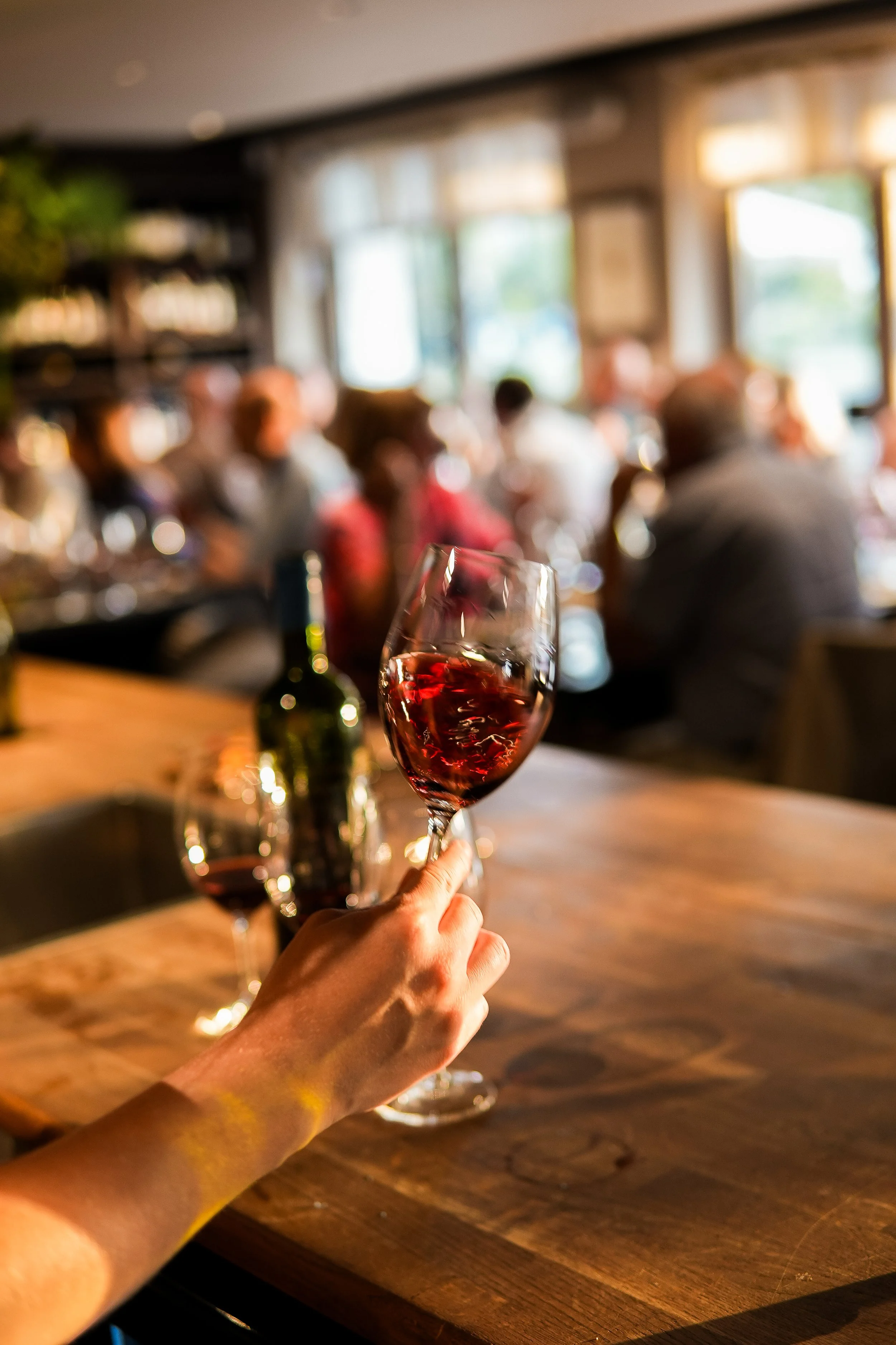 A hand holding a glass of red wine in a lively restaurant with people dining and talking in the background.