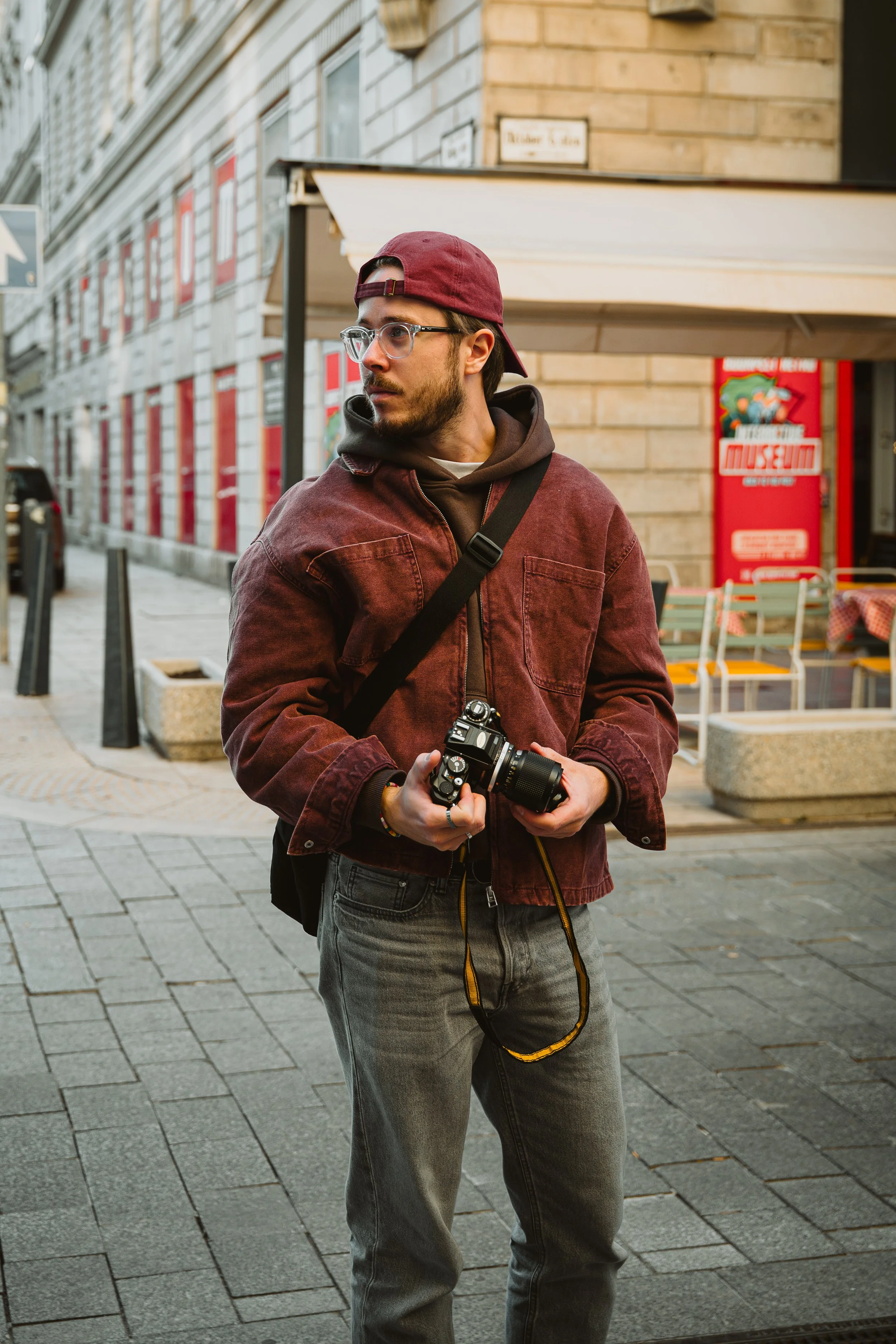 A young man with glasses, a maroon cap, and a maroon jacket holding a camera on a city street.