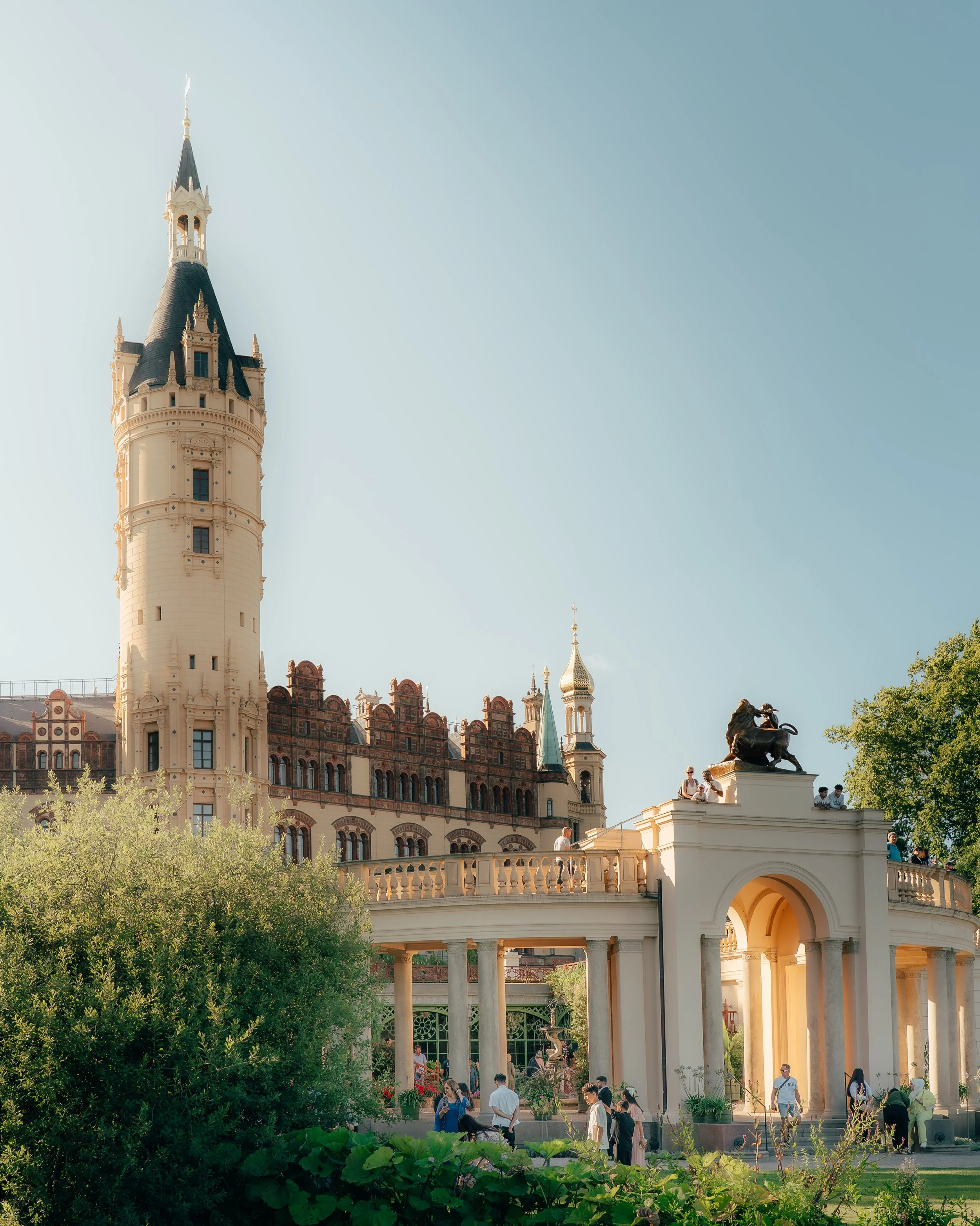 People gather near a castle with a tall central tower and a bridge adorned with statues of lions, set against a clear blue sky.