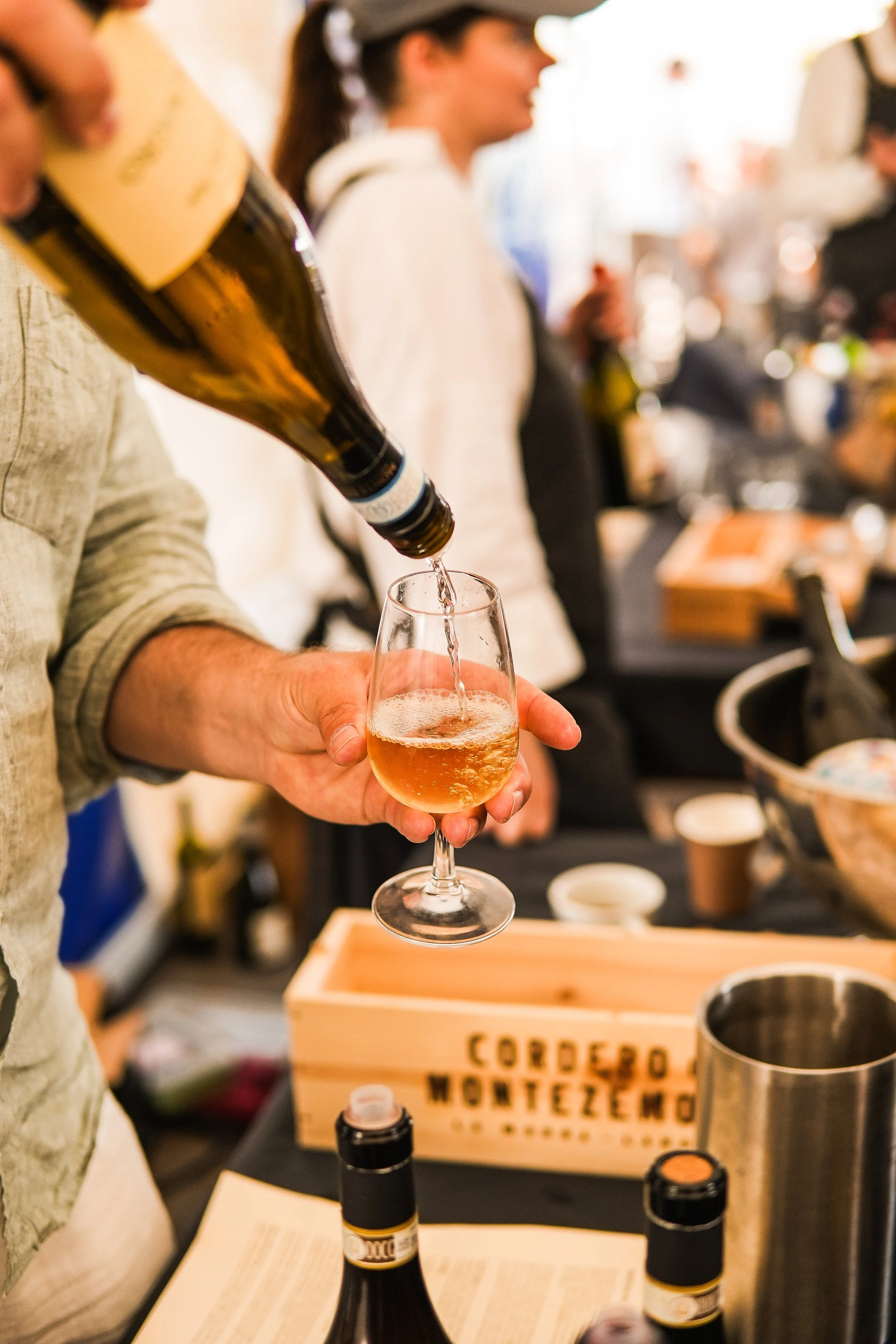 A person pouring a glass of rosé wine at a tasting event, with other people and wine bottles in the background.