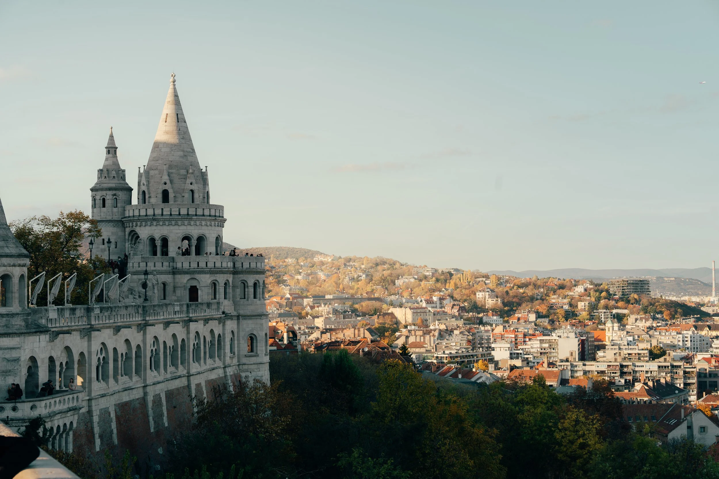 A scenic view of a European cityscape with the Fisherman's Bastion and the Danube River in Budapest, Hungary. The architecture features turrets and arches, overlooking residential and commercial buildings with rolling hills in the background under a 