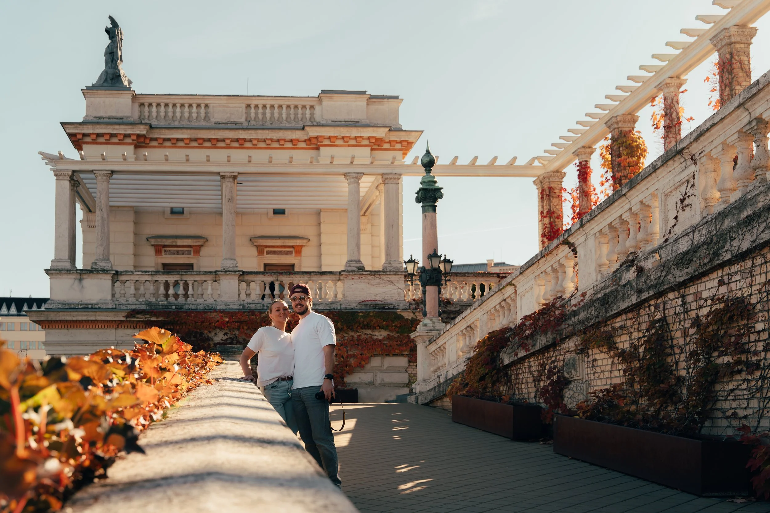 A couple posing on a city terrace with historical architecture and autumn foliage in the background.