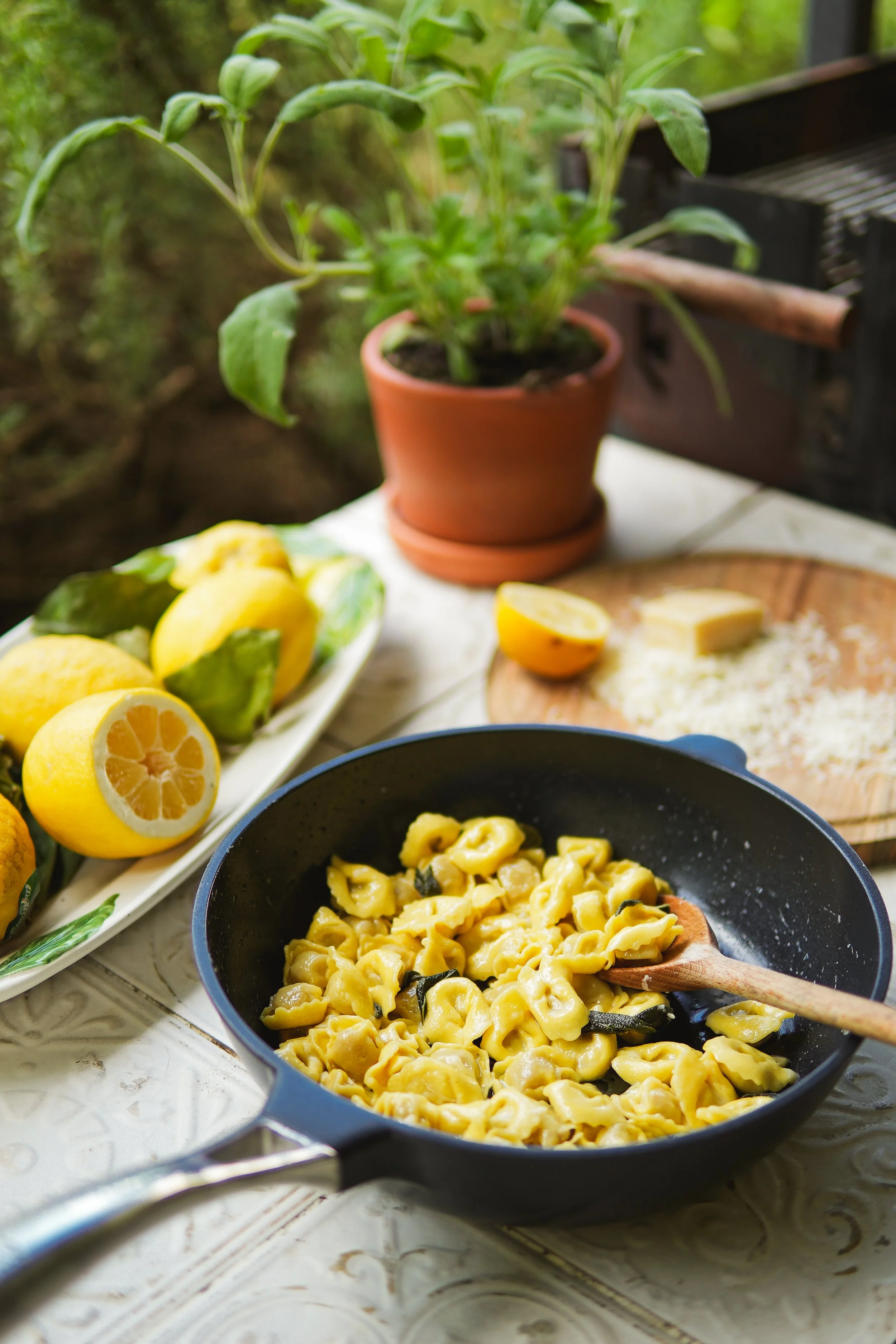 A frying pan with cooked tortellini pasta with herbs, a plate of fresh lemons, a halved lemon, and grated cheese on a wooden cutting board, with a potted basil plant in the background.