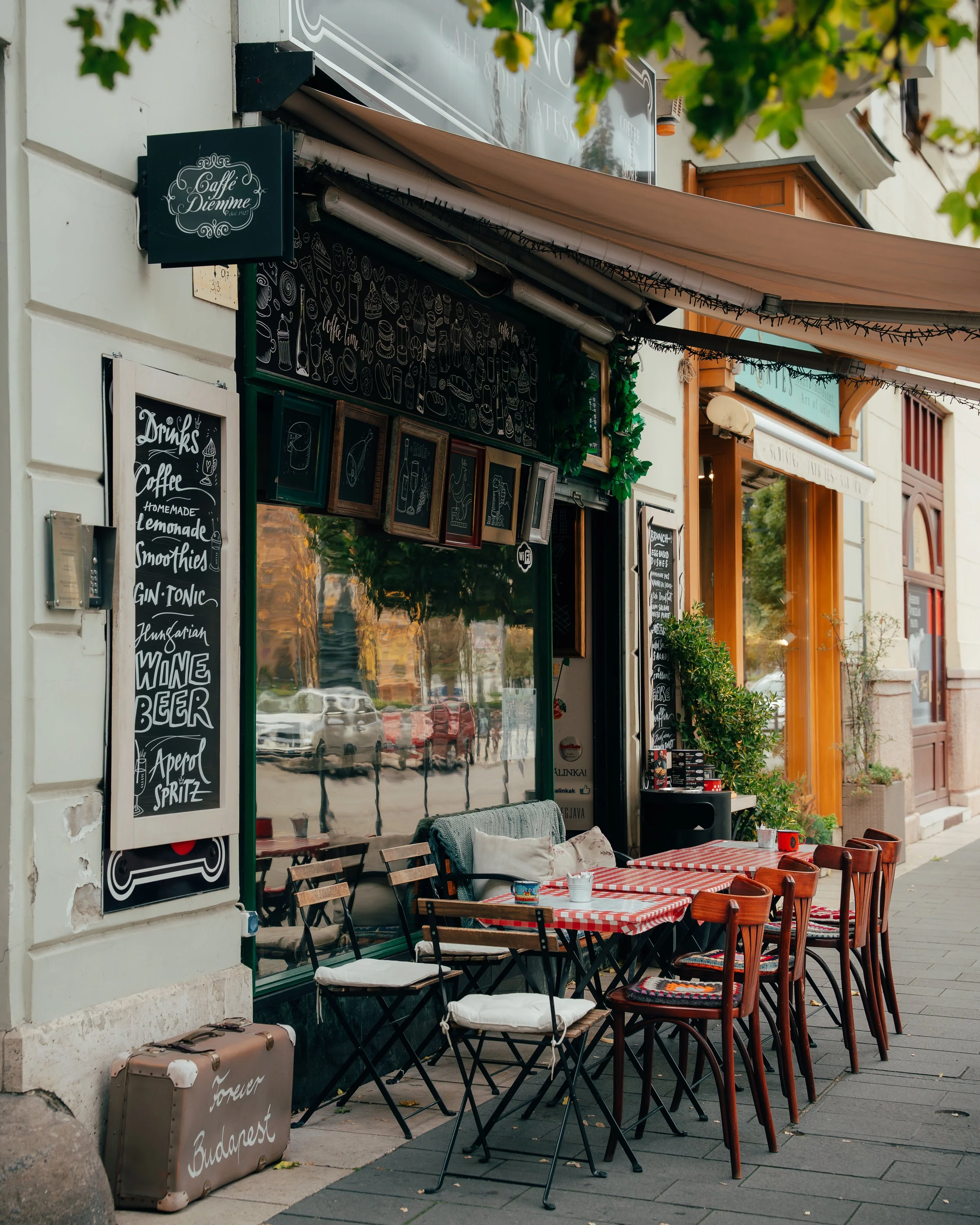 An outdoor cafe with red checkered tablecloths and wooden chairs, some with white cushions, situated on a city sidewalk. The cafe has black menu boards with white writing and framed chalkboard art. A suitcase with 'Forever Budapest' written on it is 