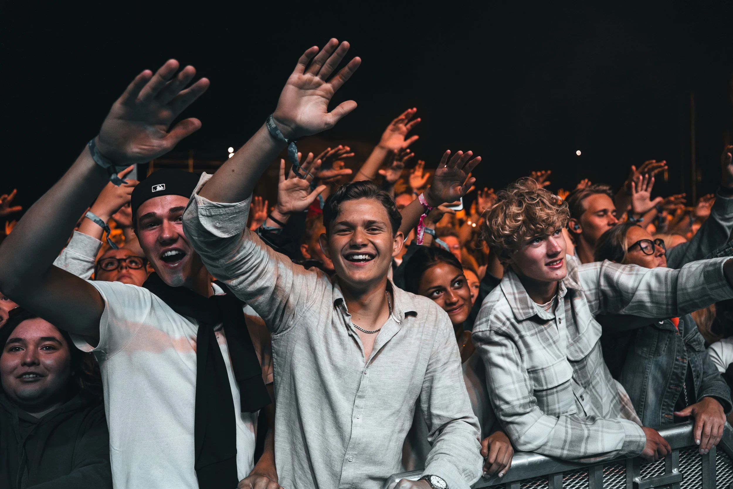 Crowd of young people at a concert or event, cheering and smiling, some with hands raised, at night.