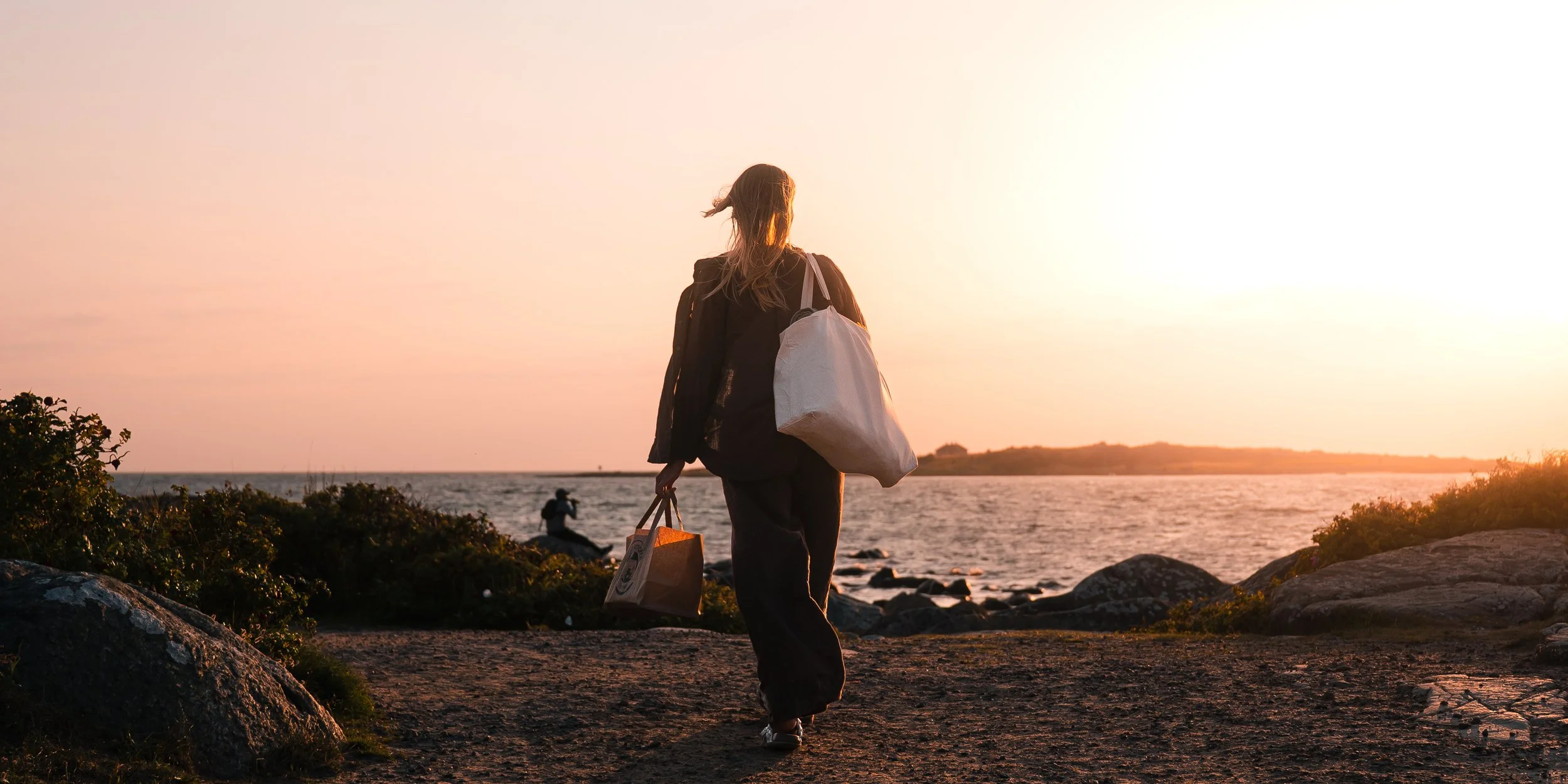 A woman walking along a rocky beach at sunset, carrying shopping bags.
