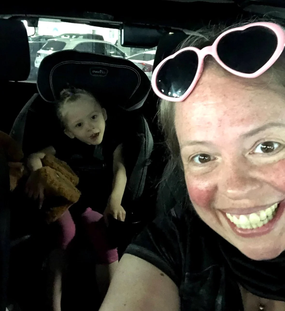 A picture of a woman and child smiling in a car.  The woman has pink sunglasses on her forehead, and the child is in a car seat holding a large stuffed bear.