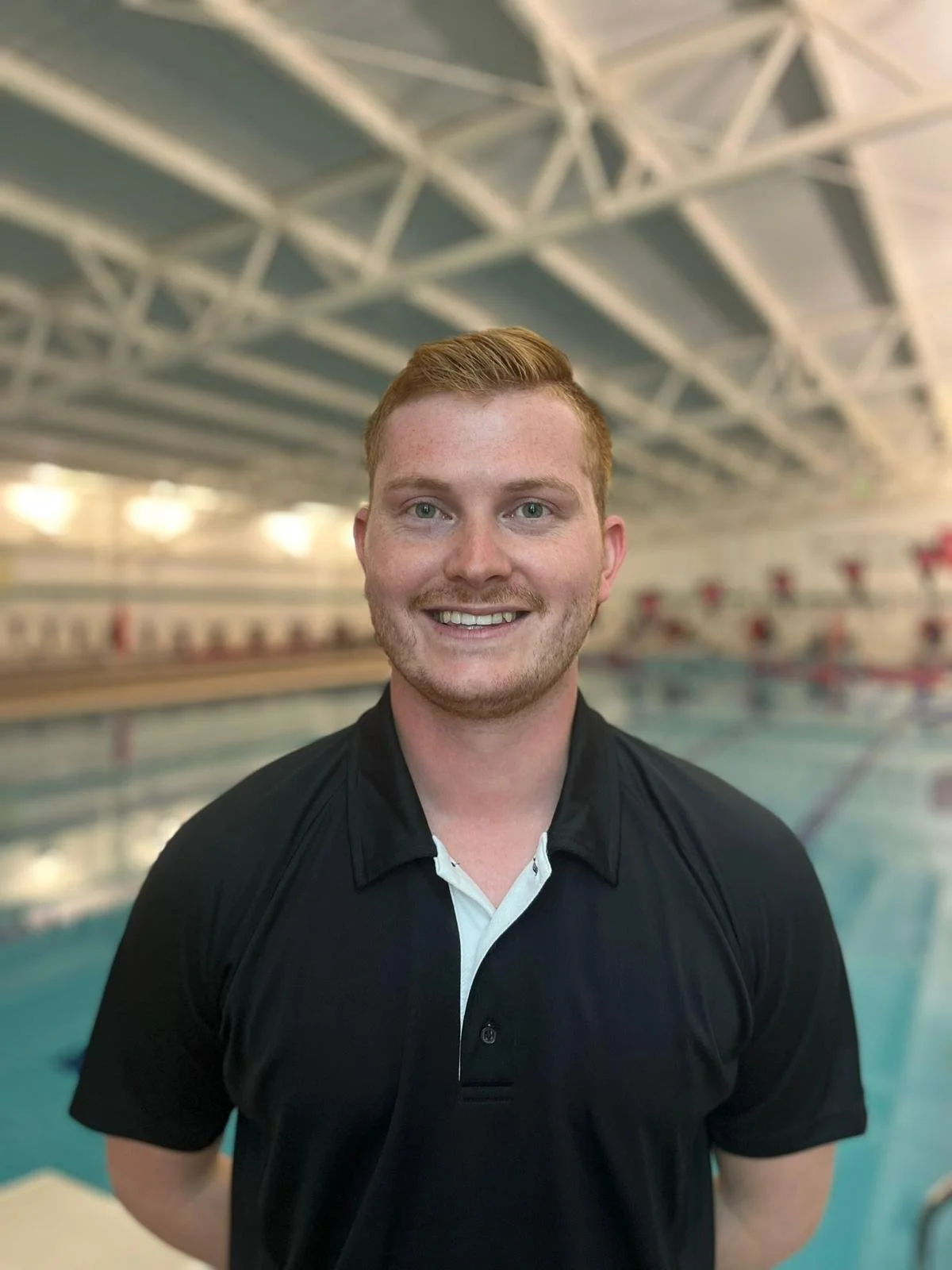 A young man with red hair and a beard smiling in front of an indoor swimming pool, wearing a black and white polo shirt.