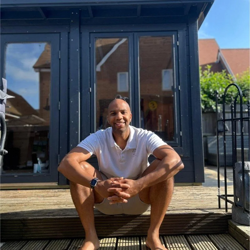 A smiling man sitting cross-legged on a wooden deck outside a black-framed glass house. He is wearing a white polo shirt and shorts, with a smartwatch on his left wrist. The background includes parts of a backyard with green trees and neighboring houses.