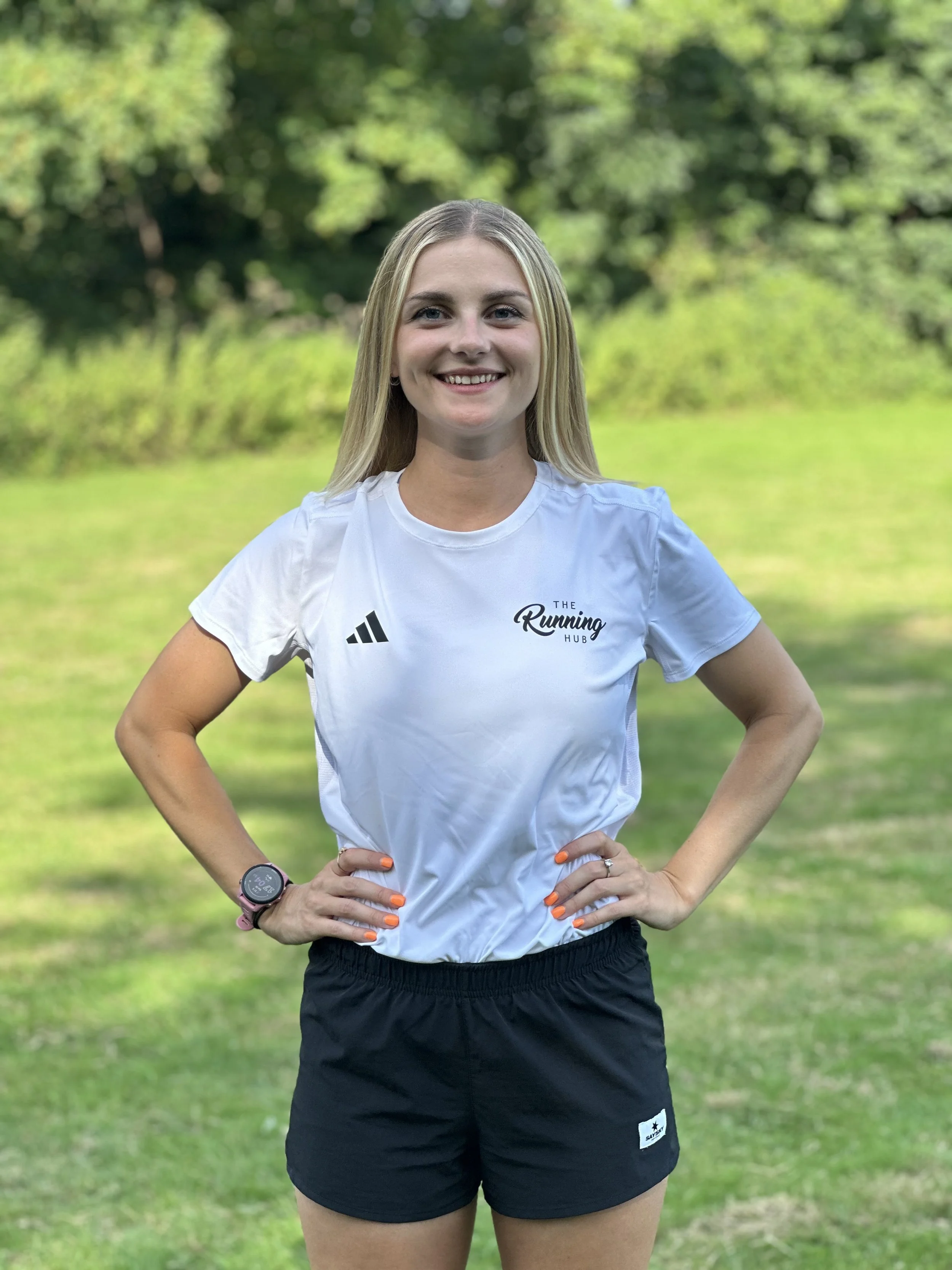A woman with long blonde hair standing outdoors on grass with green trees in the background. She is smiling, wearing a white athletic t-shirt with black shorts, a watch, and has orange-painted nails.