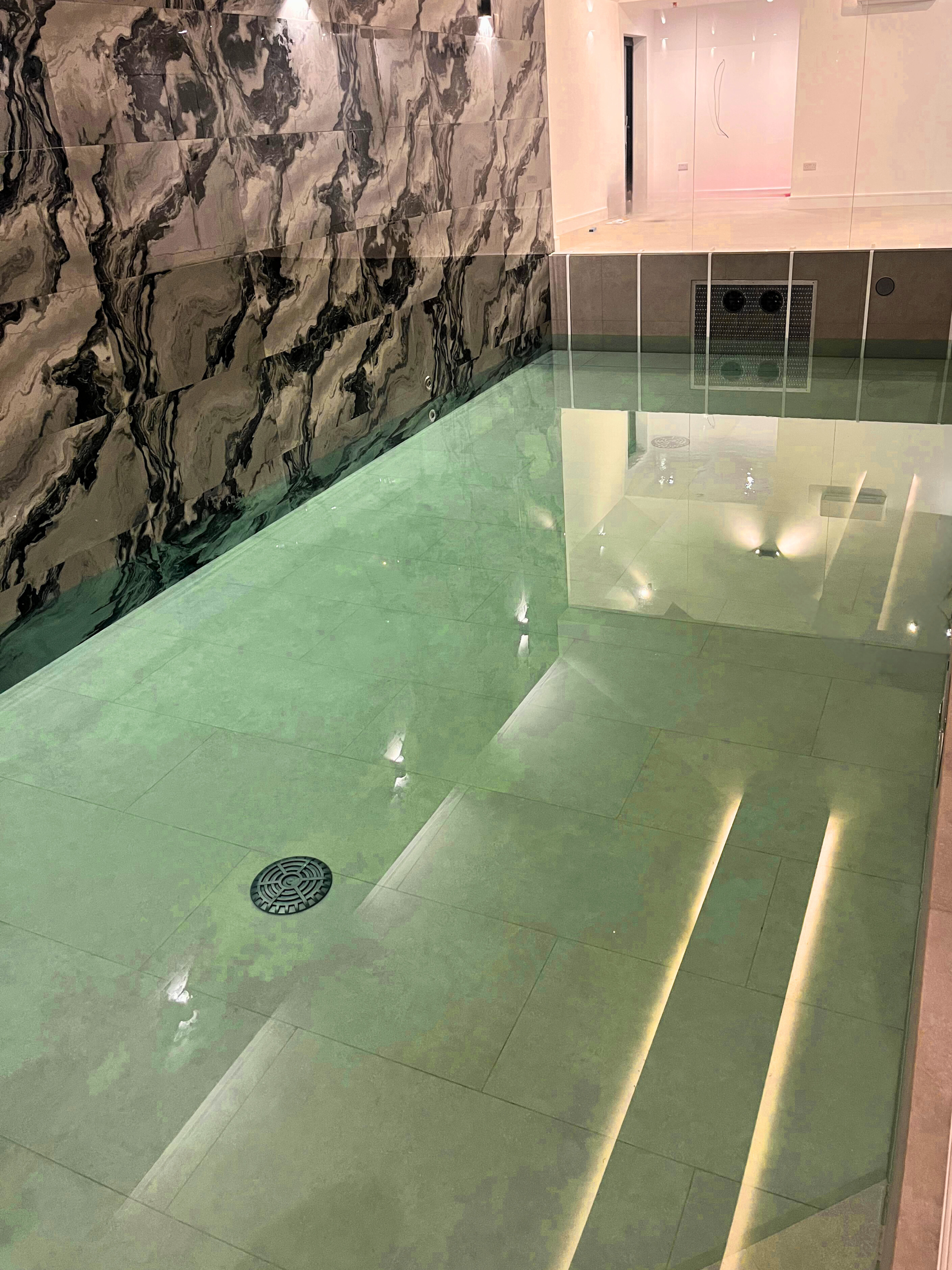 Indoor swimming pool with green water, marble wall, and glass divider, reflecting ceiling lights.