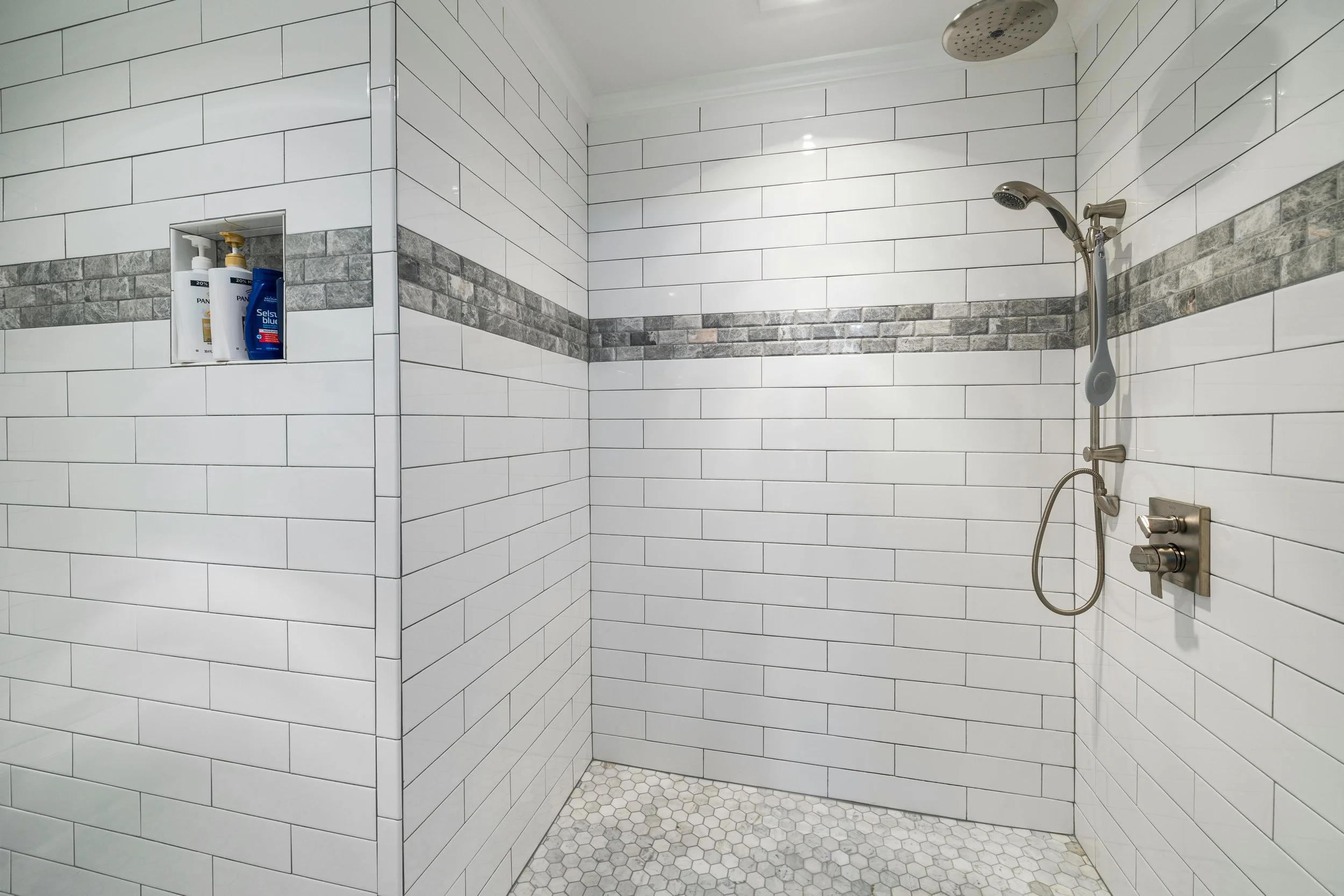 Empty walk-in shower with white subway tile walls, a horizontal strip of gray tile accents, a built-in niche with toiletries, and a shower with a handheld and rain showerhead.