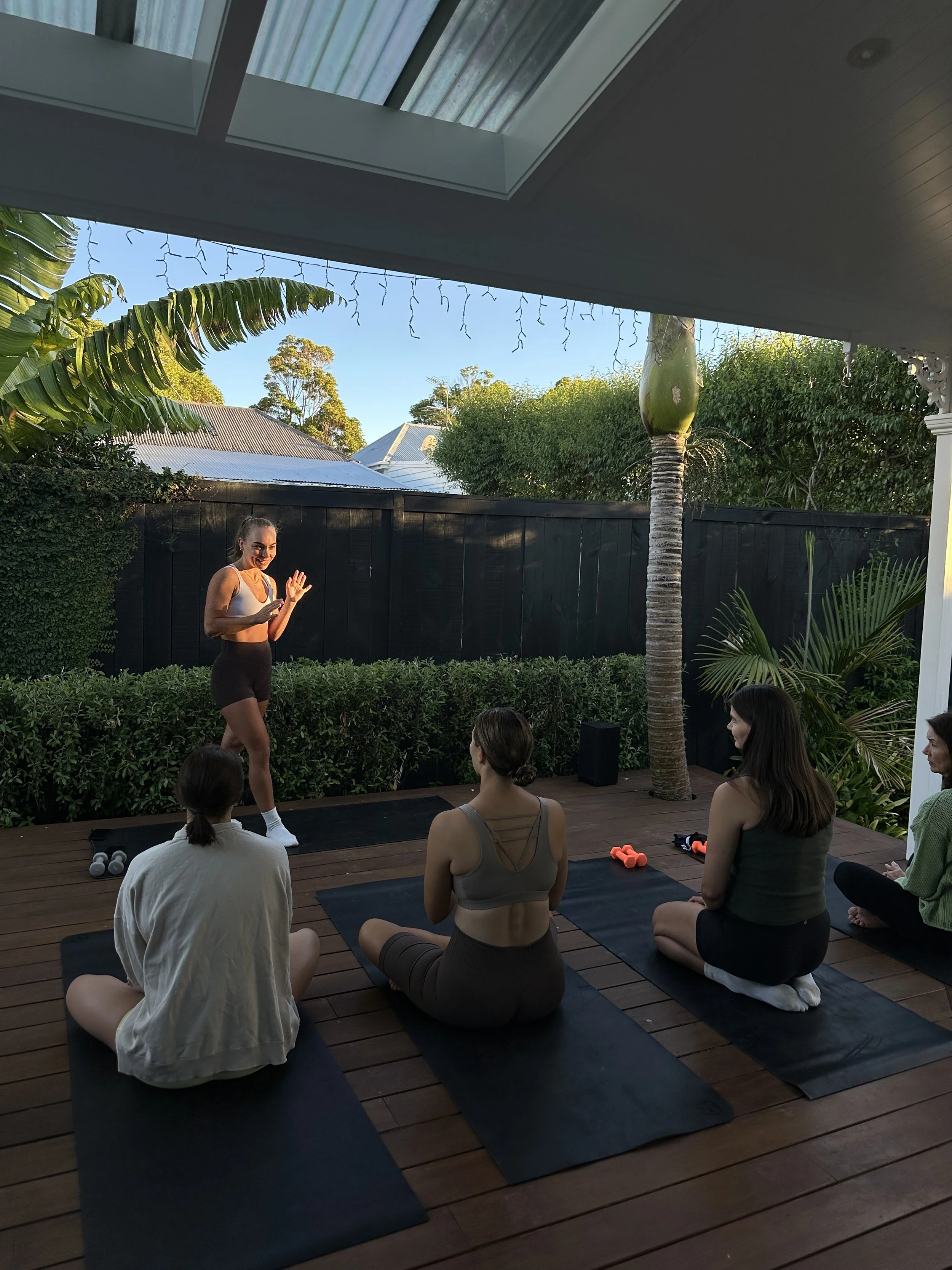 A woman leading a yoga class on a wooden patio outdoors, with four women sitting on yoga mats, facing her and practicing yoga poses in the late afternoon sun.
