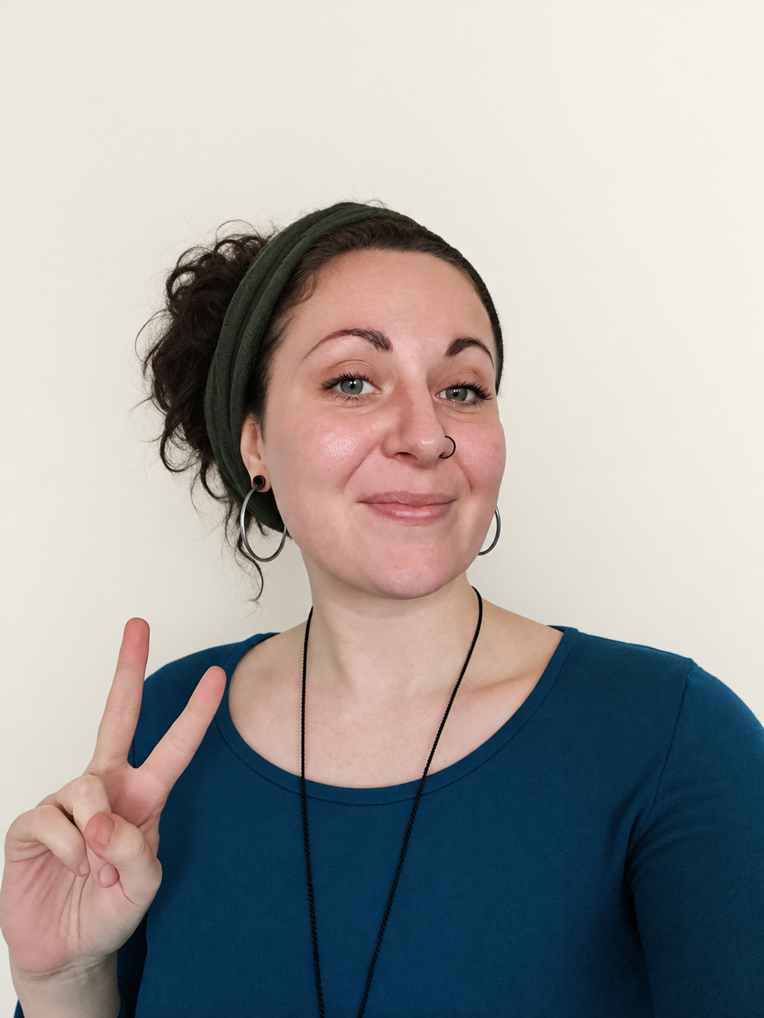 Hannah, a web designer based in Ely, Cambridgeshire, smiles at the camera while throwing a peace sign. She has a dark blue top, a dark green headband and silver hoop earrings.