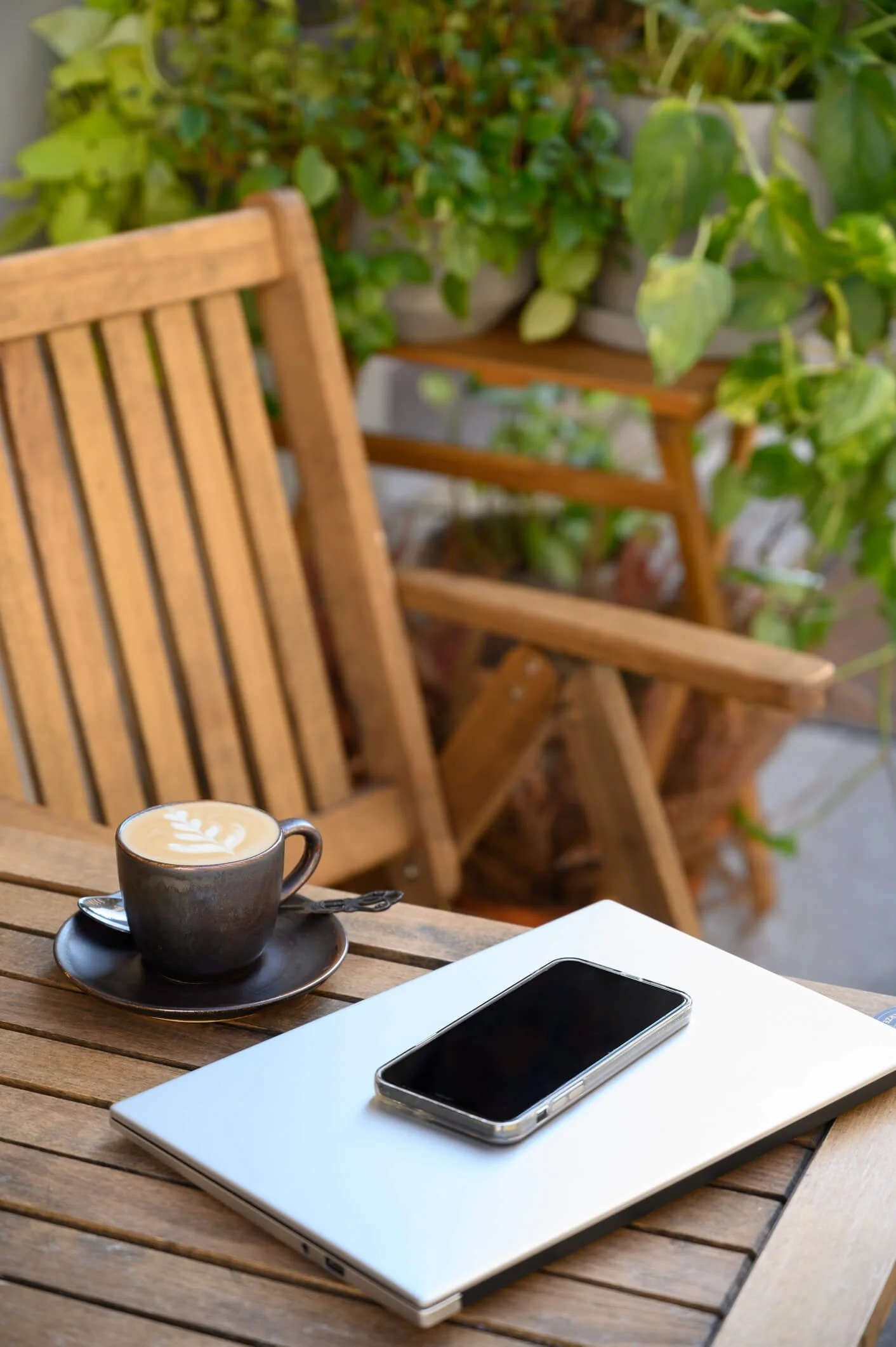 A work laptop sits on a wooden table outdoors at a cafe. There is a mobile phone and a mug of coffee on the table, a chair pulled out, and leafy, green plants in the background.