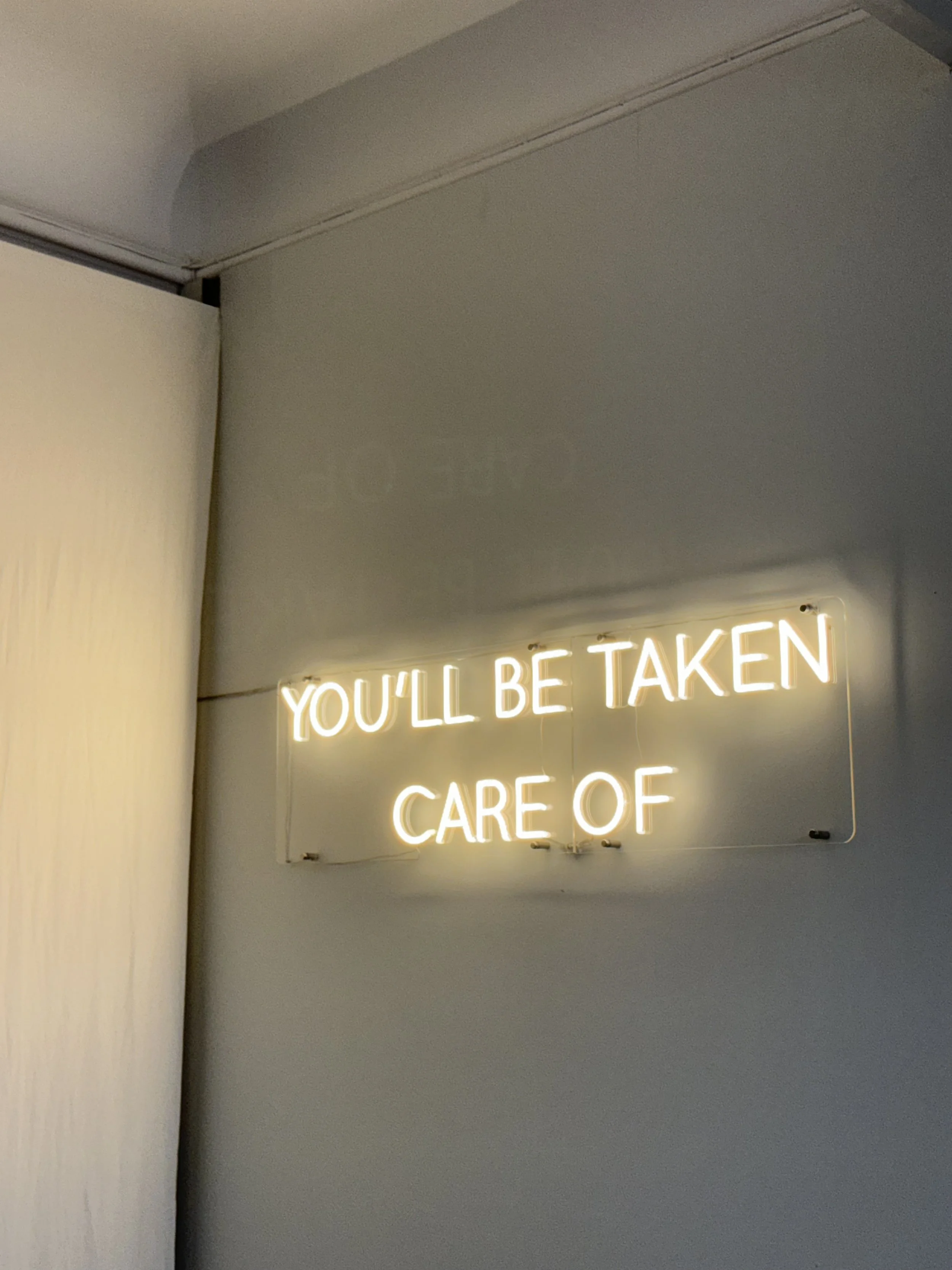Neon sign with white glowing letters saying, 'YOU'LL BE TAKEN CARE OF,' mounted on a wall.
