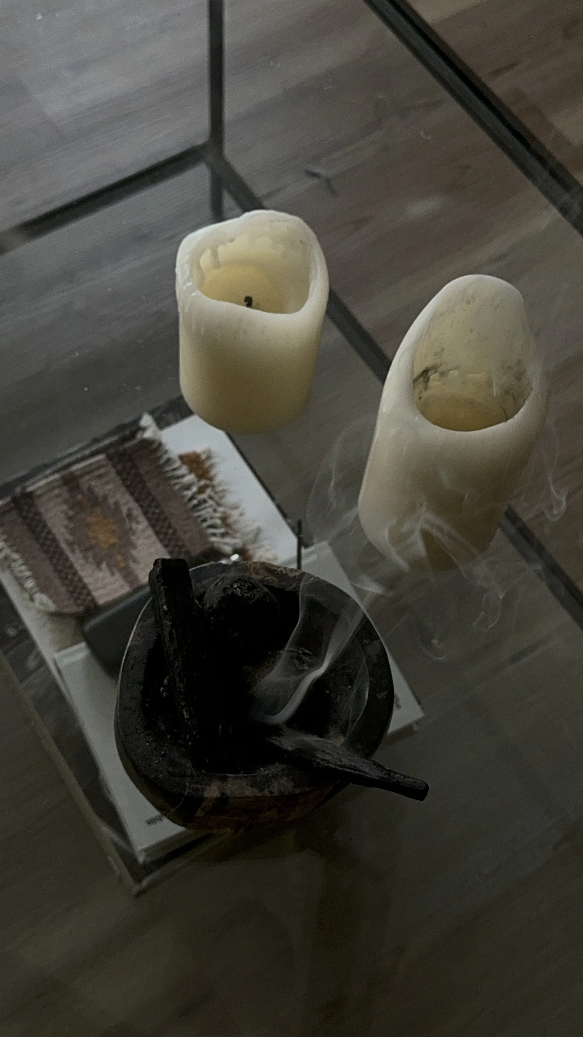 Two white candles and one dark candle with melted wax and smoke standing on a glass table, with a woven rug and a box in the background.