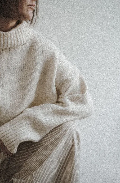 Close-up of a person wearing a cream-colored knit turtleneck sweater and beige corduroy pants, sitting against a plain background.