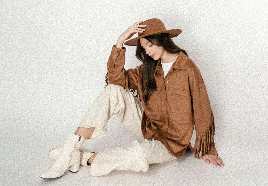 Young woman sitting and leaning on the floor, wearing a brown hat, brown fringed jacket, cream pants, and white boots, against a plain light background.