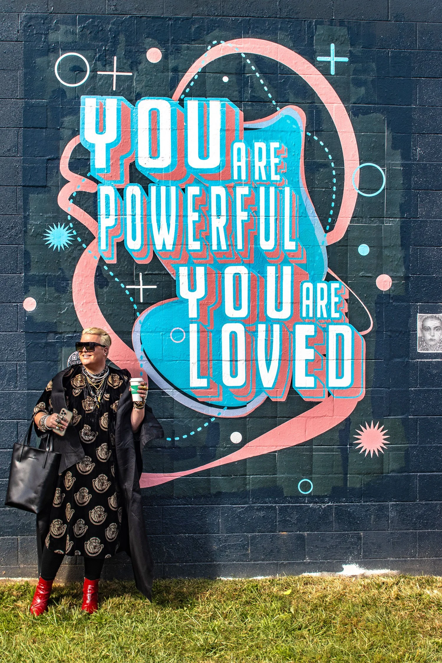 A woman with blonde hair and sunglasses stands in front of a colorful mural on a brick wall that reads 'You are powerful. You are loved.' She holds a coffee cup in one hand and a phone in the other and is wearing a black dress with a patterned coat, jewelry, and red boots.