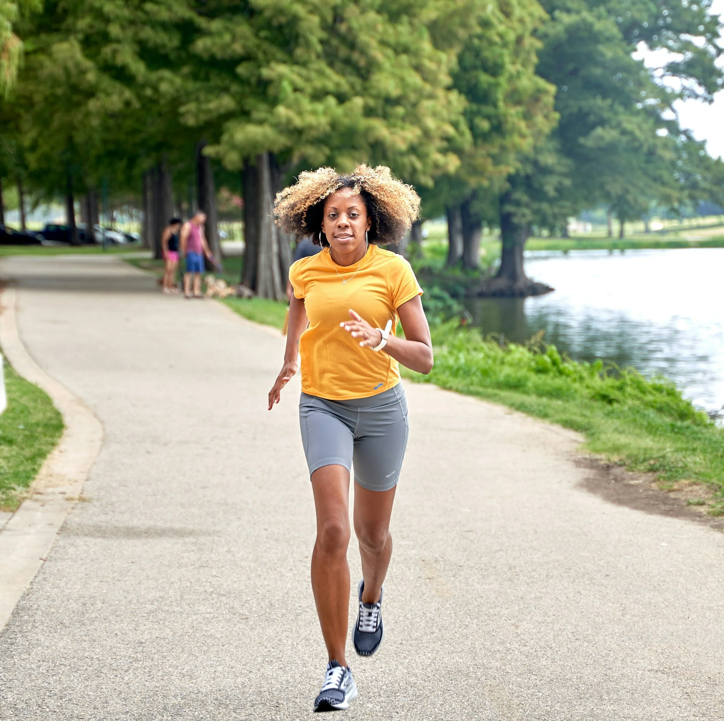A woman jogging on a paved pathway beside a lake in a park with trees in the background.