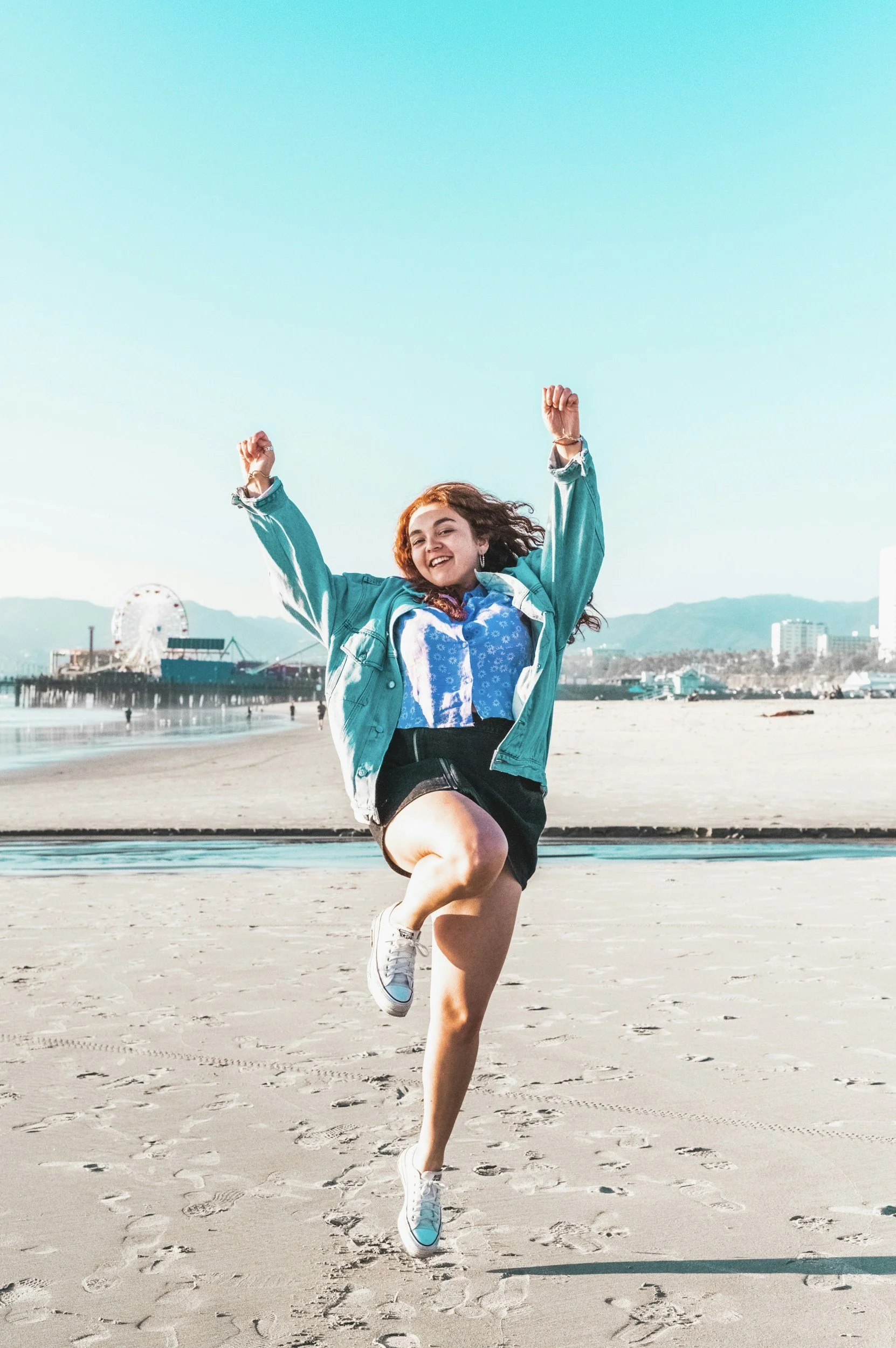 A woman joyfully jumping on a beach with a Ferris wheel and cityscape in the background on a sunny day.