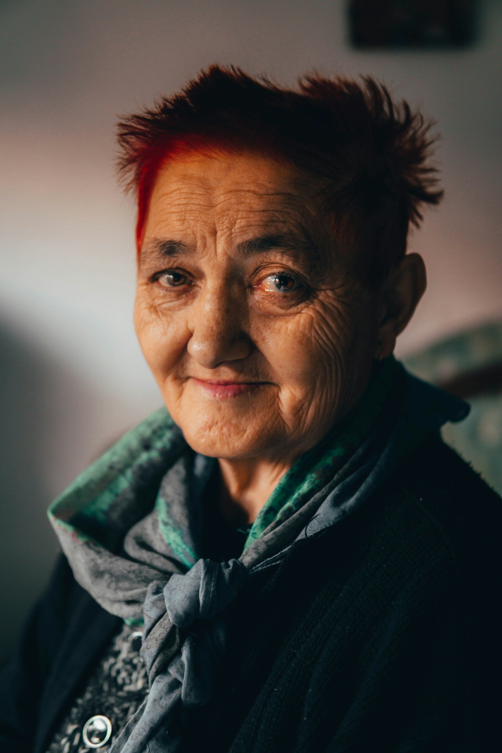 An elderly woman with short, dark, slightly red-tinted hair and blue eyes, wearing a patterned scarf around her neck, smiling softly in a close-up portrait.